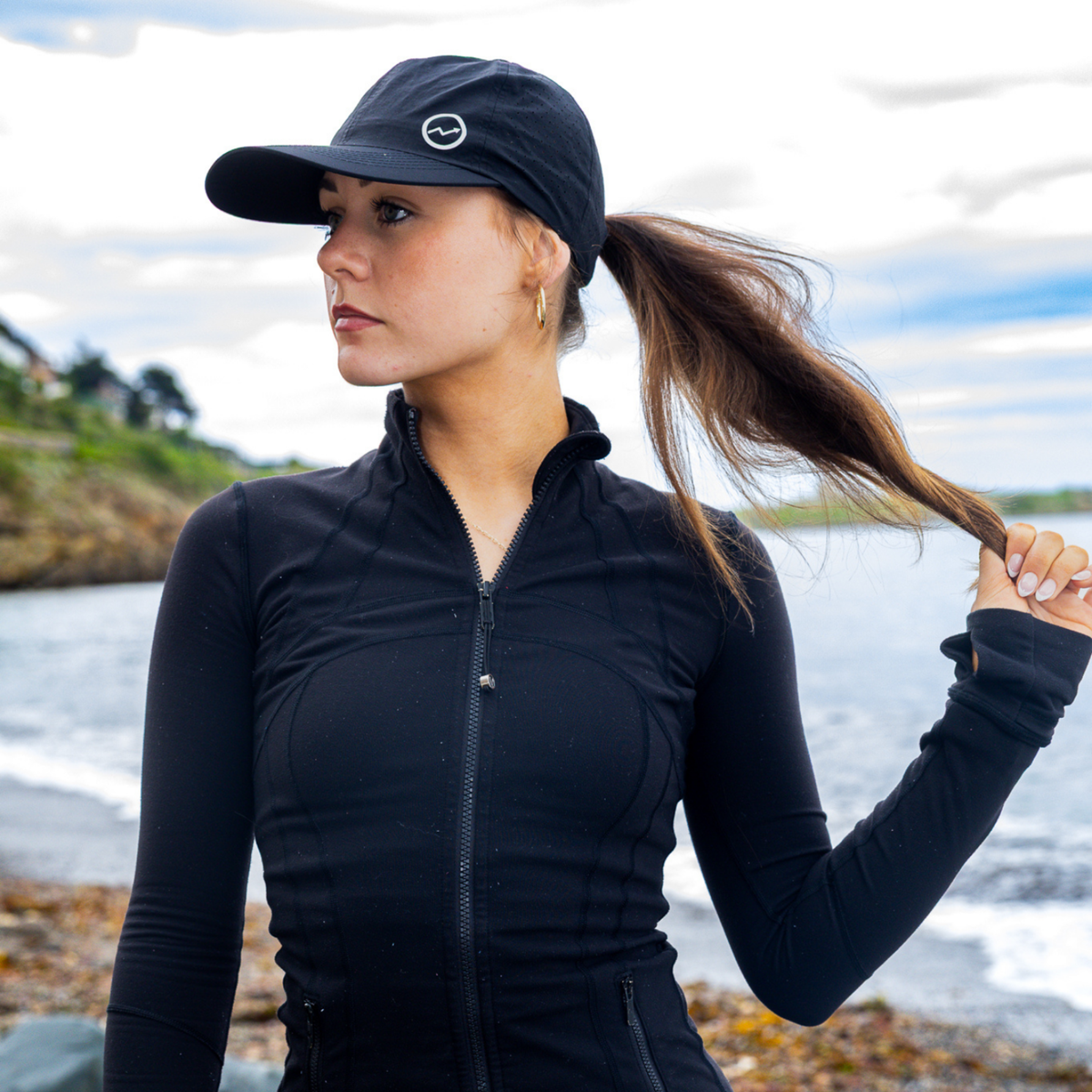 A woman wearing a black long-sleeve athletic zip-up jacket and a Eos Elite Unisex HighER Running Cap Black EOS-CAP1 stands on a rocky shoreline with water and cloudy sky in the background.