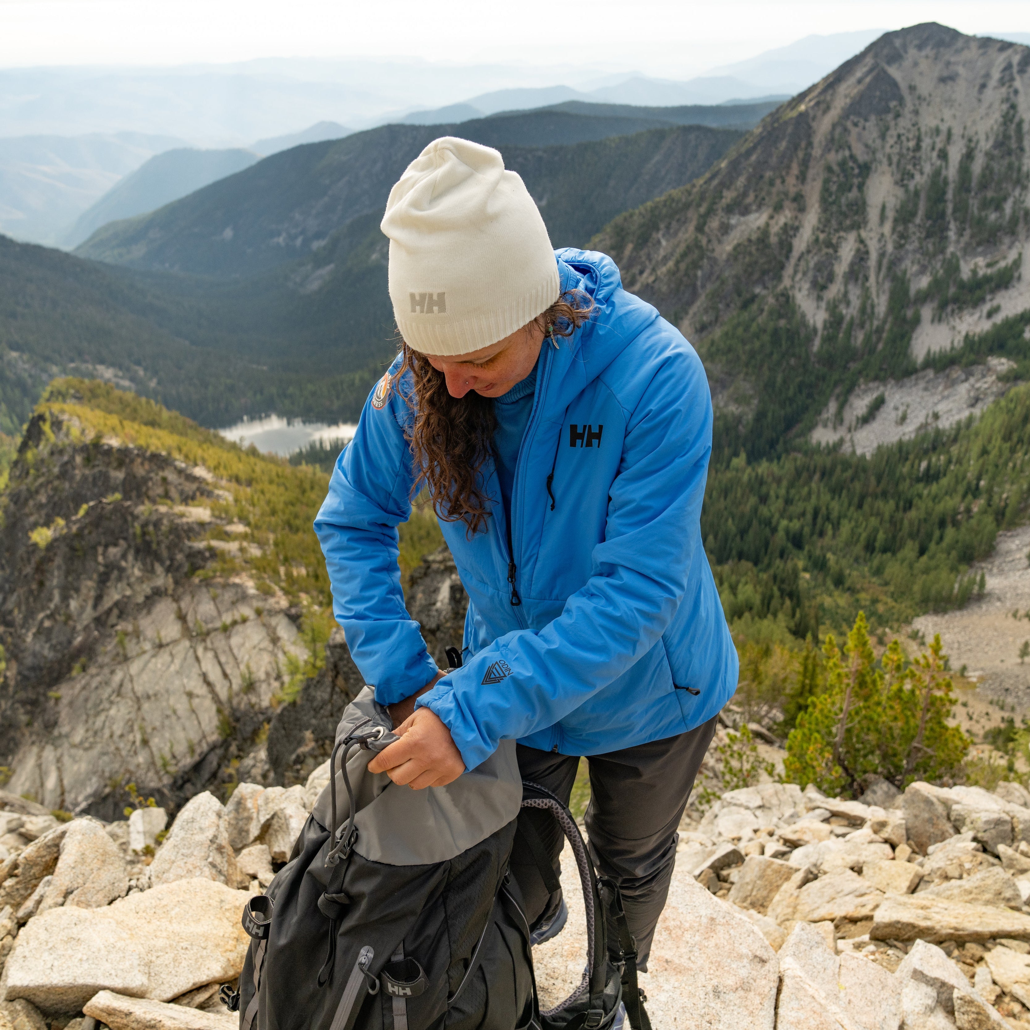 Woman wearing Helly Hansen Unisex Brand Beanie Hat White 001 57502 and blue Helly Hansen jacket on the top of a mountain
