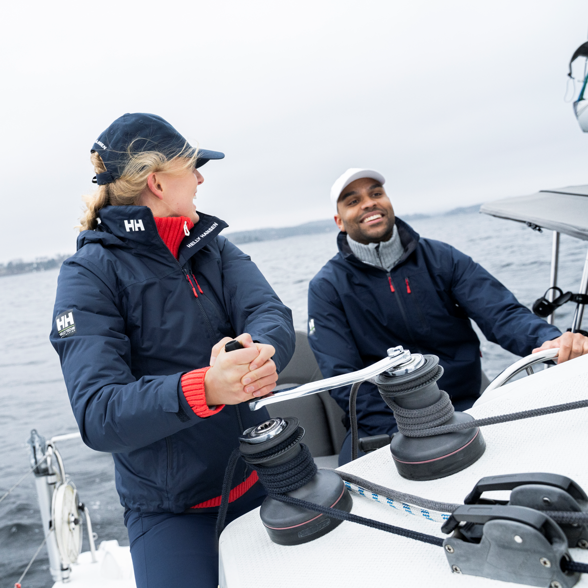 Two people on a sailboat in a body of water. Woman wearing Helly Hansen Women's Crew Hooded Midlayer Waterproof Insulated Jacket 2.0 Navy 597 34447
