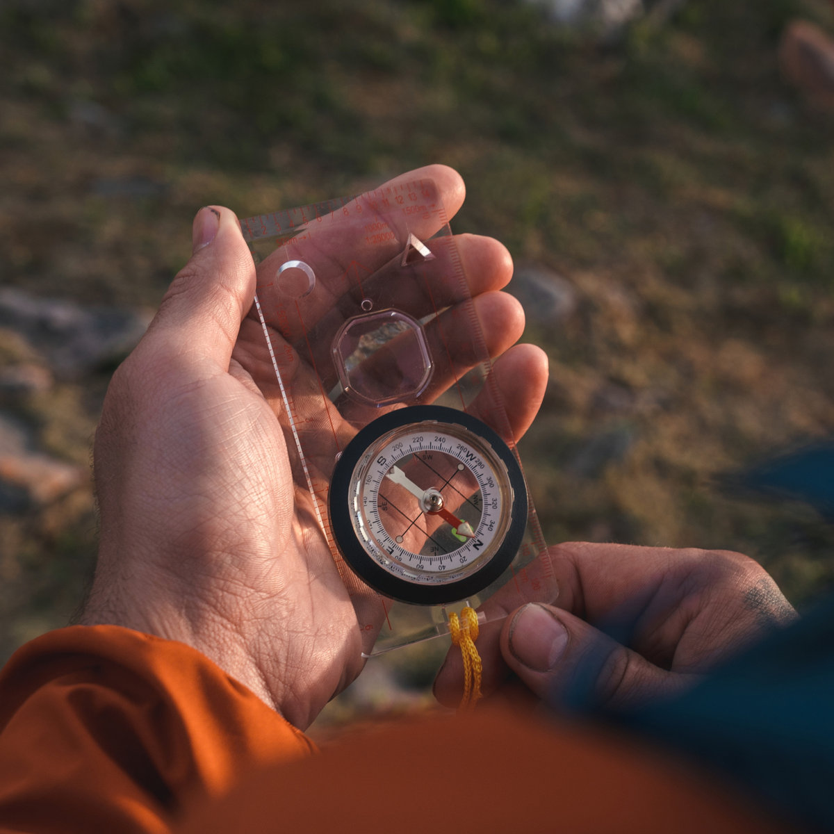 A person holds a Highlander Deluxe Map Compass COM006, with blurred natural ground in the background.