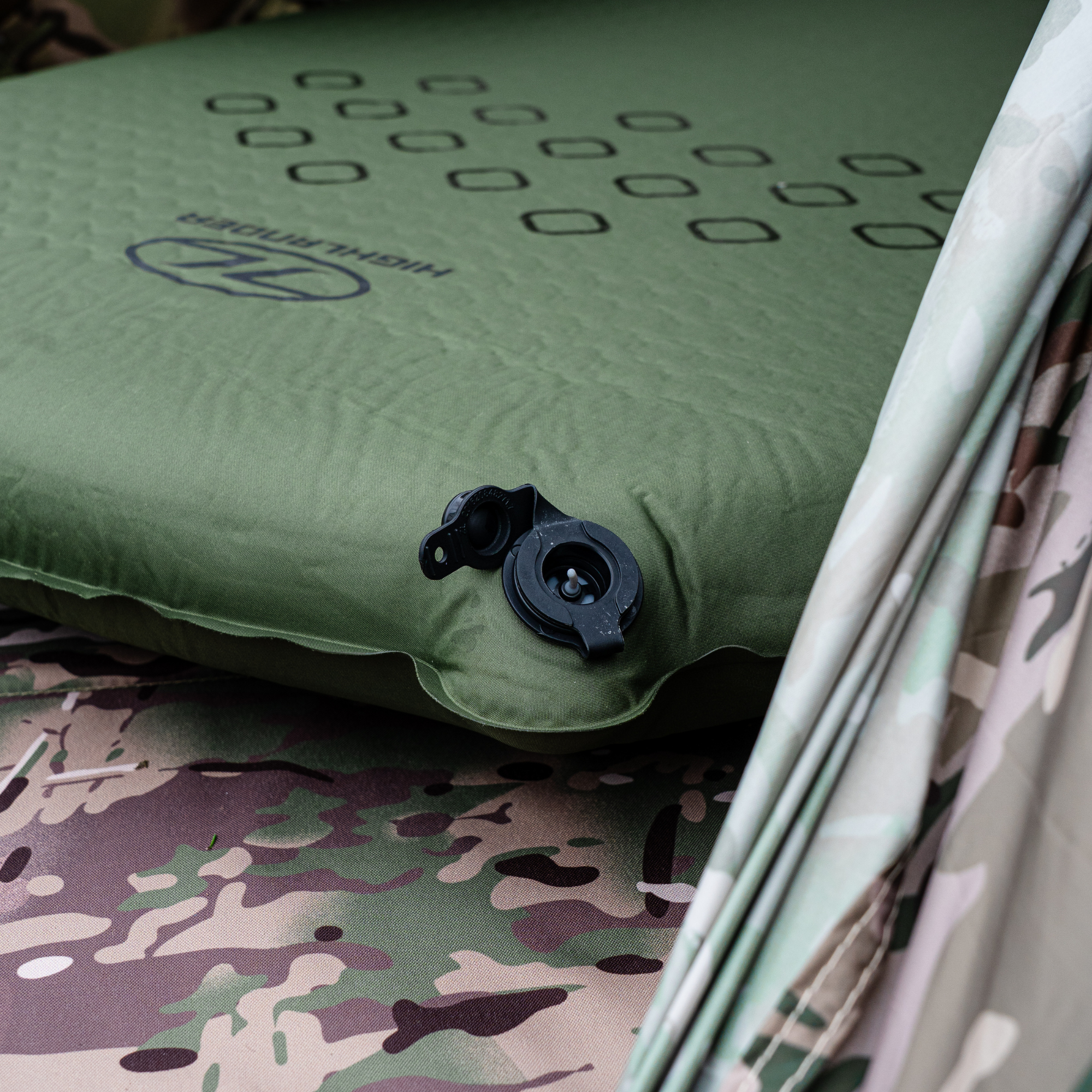 Highlander Self Inflating Kip Sleeping Mat 75mm Olive Green SM132-75 shown resting on a camouflage-patterned surface next to a matching rolled sleeping bag or fabric.