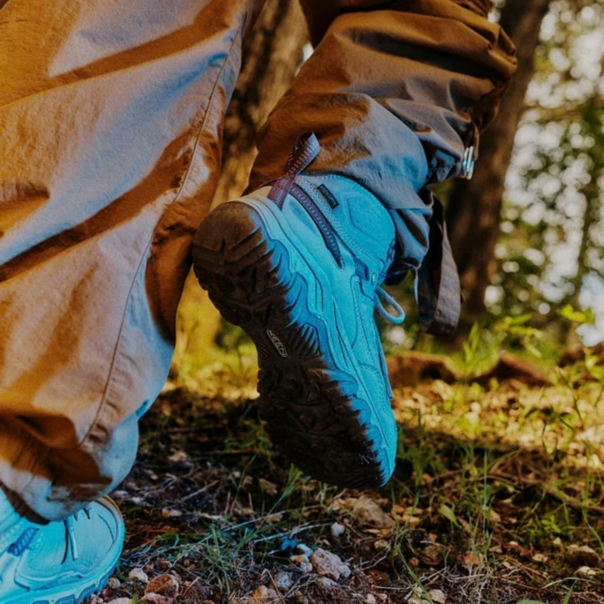 A close-up outdoor shot of a person hiking, wearing Keen Men's Targhee IV Waterproof Mid Trail Boots Magnet Black 1032633, paired with brown trousers in a forest setting.