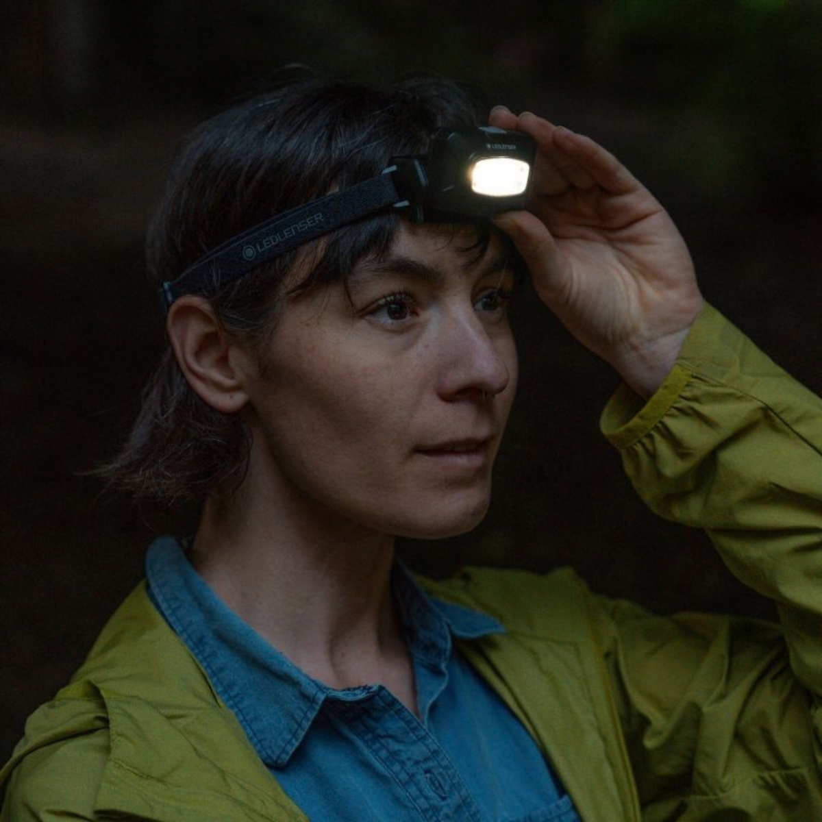 A woman outdoors wearing a Ledlenser HF4R CORE Rechargeable Headlamp Black 502790, adjusting the lamp. She is wearing an olive green jacket over a blue shirt, with a dark, shadowy forest background.