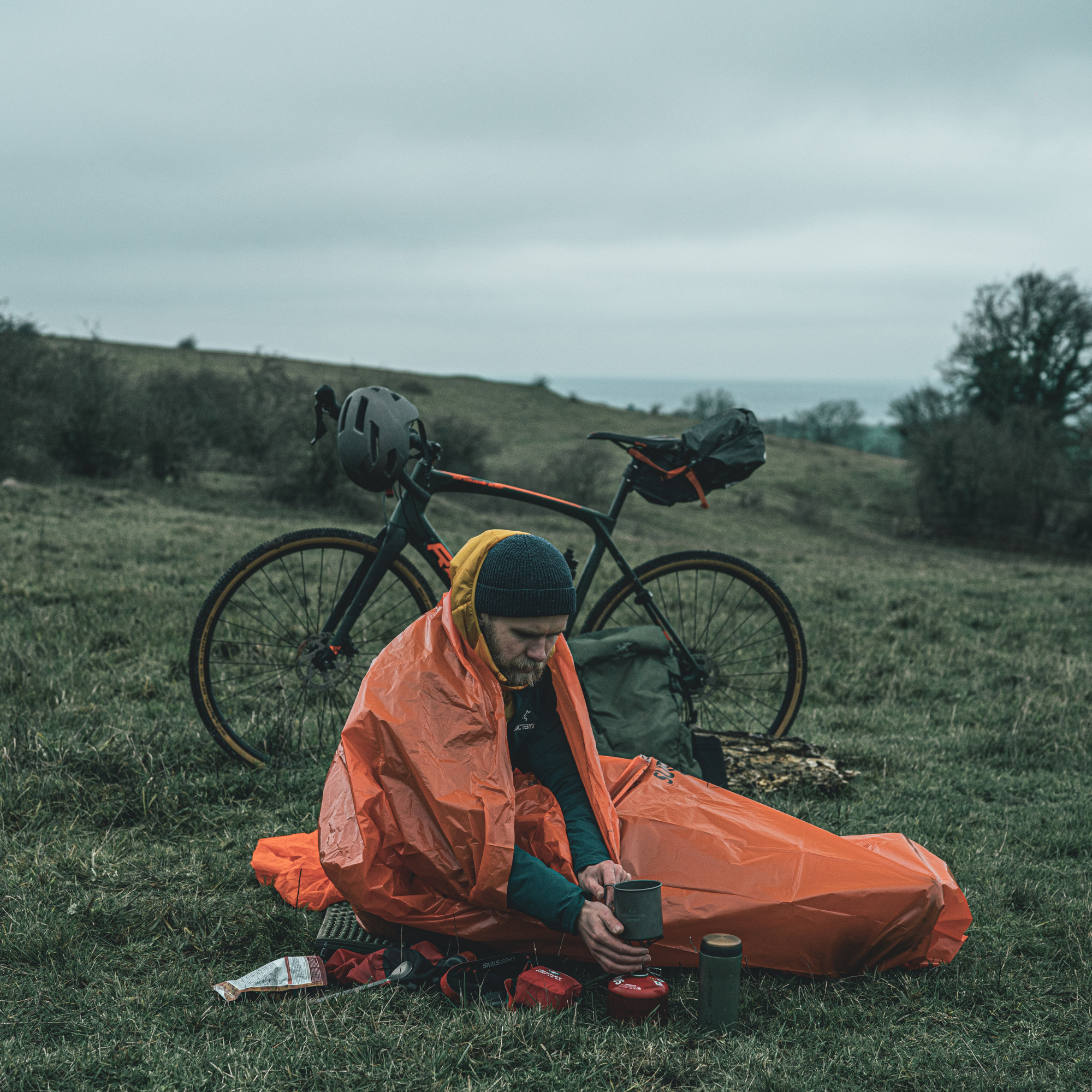 A person sitting on grass in an outdoor camp setting, wrapped in a Lifesystem Emergency Survival Bag Orange 2090, with a bicycle and camping gear nearby under an overcast sky.