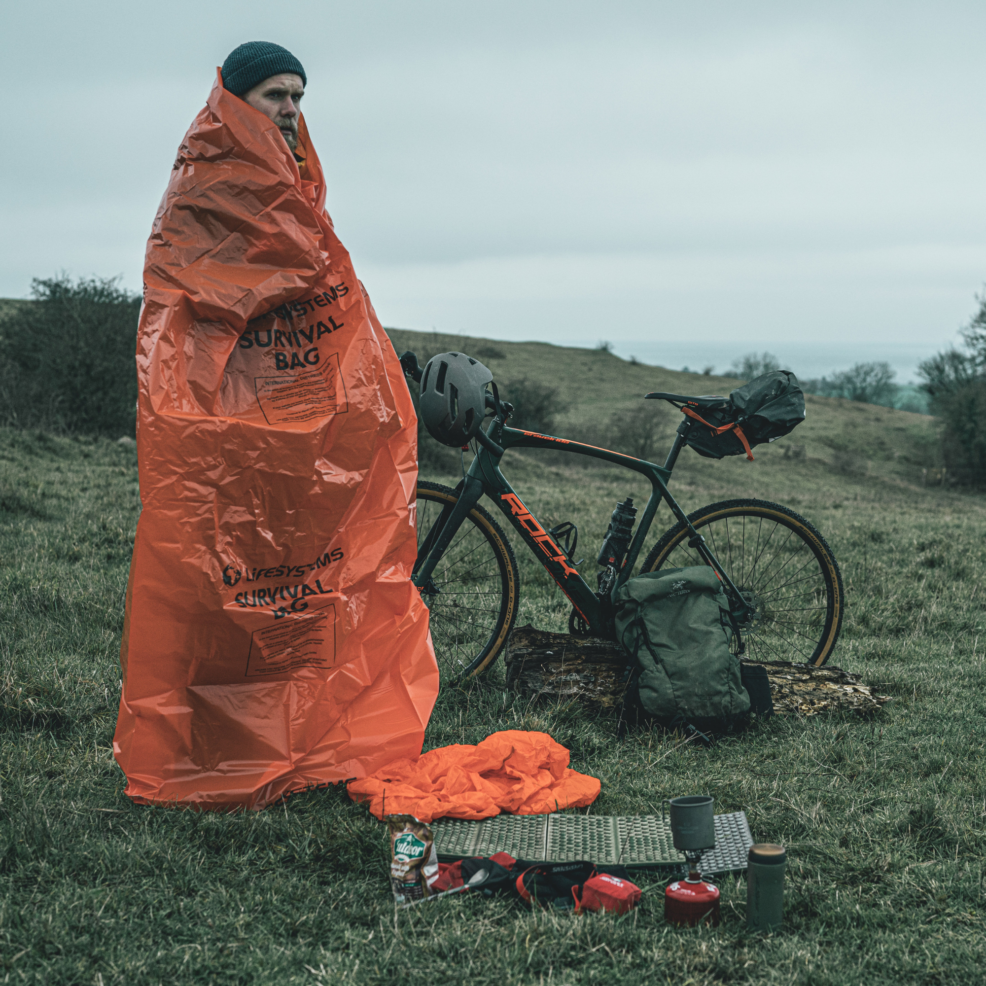 A person standing outdoors in a grassy field wrapped in a Lifesystem Emergency Survival Bag Orange 2090, with a bicycle and camping gear nearby under an overcast sky.