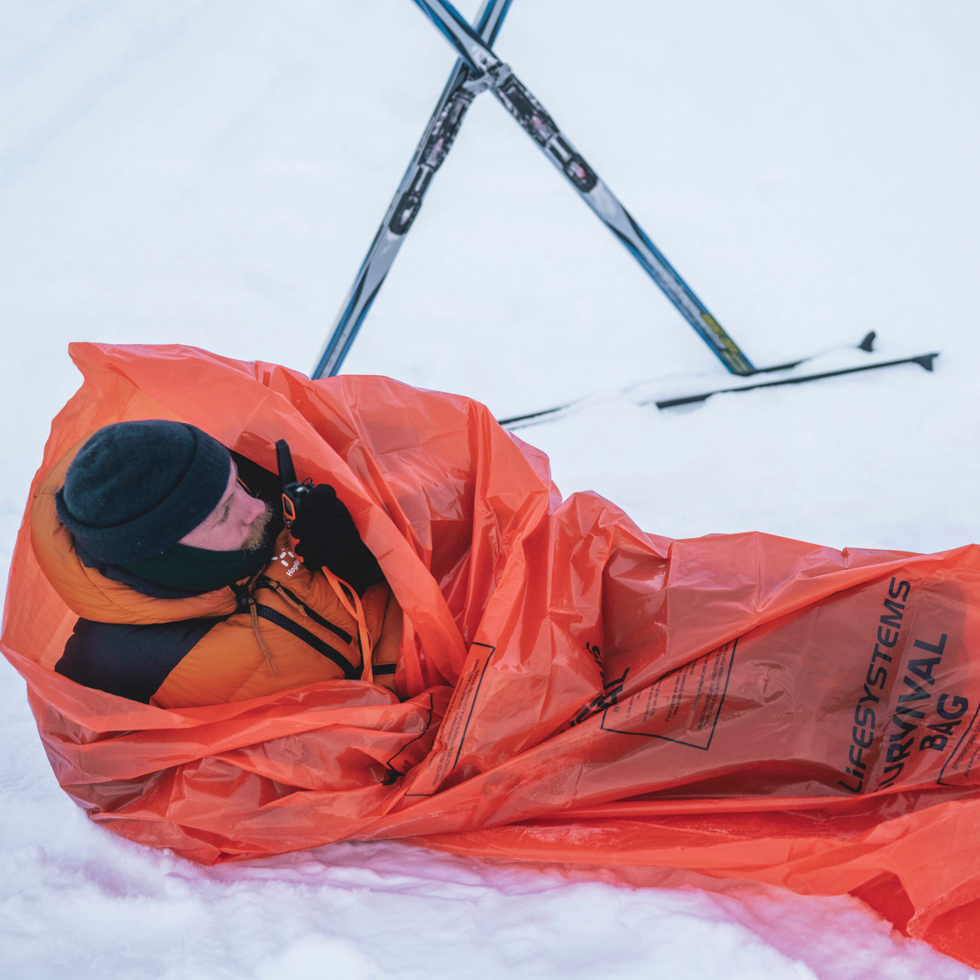 A person lying in deep snow wrapped in a Lifesystem Emergency Survival Bag Orange 2090, with skis visible in the background.