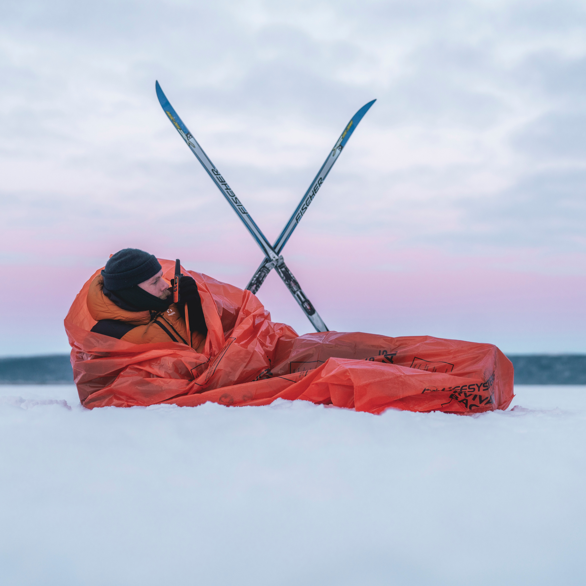 A person lying in the snow wrapped in a Lifesystem Emergency Survival Bag Orange 2090 with crossed skis behind them against a pale pink and blue sky.