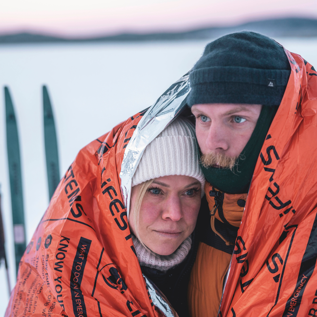 Two people in a snowy setting, with skis in the background, with a Lifesystem Heatshield Survival Blanket Double 42170 wrapped around both of them