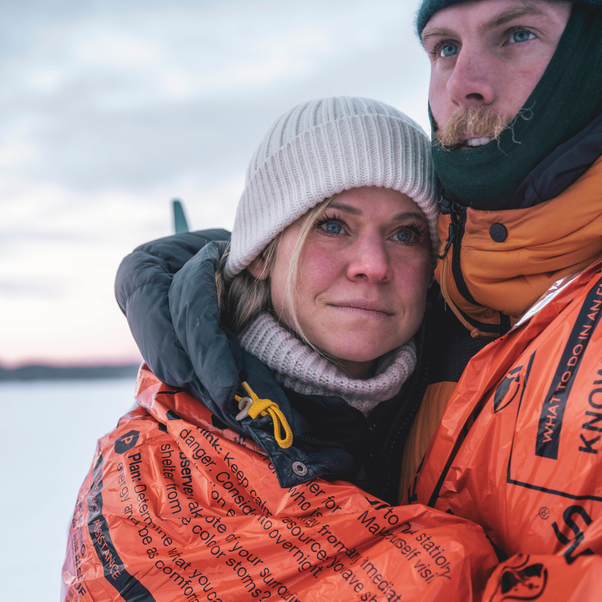 Two people in a snowy setting, with skis in the background, with a Lifesystem Heatshield Survival Blanket Double 42170 wrapped around both of them