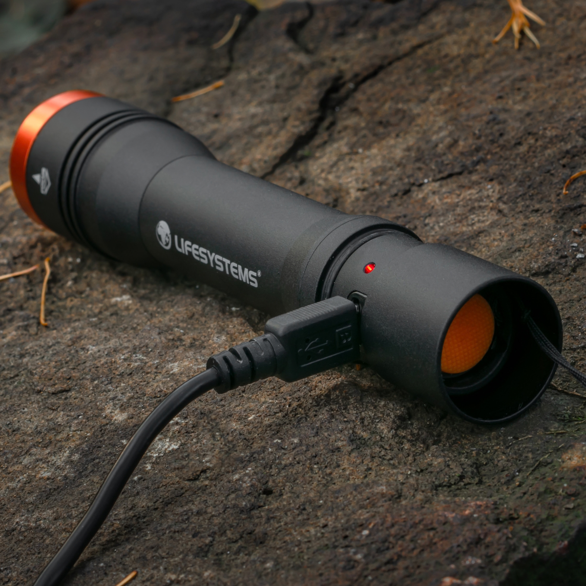 A Lifesystems Intensity 545 LED Hand Torch 42061 with orange accents, shown resting on a textured rock outdoors with a USB charging cable plugged in.