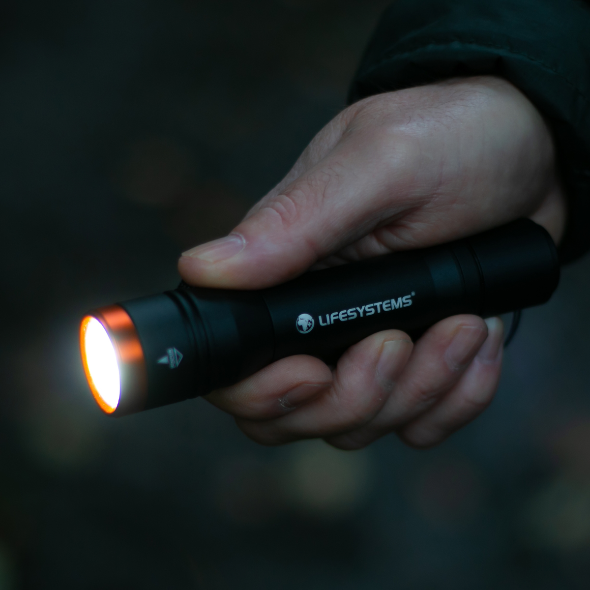 A hand holding a Lifesystems Intensity 545 LED Hand Torch 42061 with its light illuminated, set against a dark, blurred outdoor background.