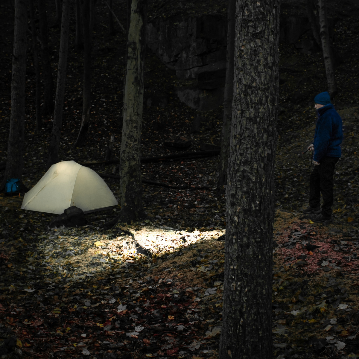 A Lifesystems Intensity 545 LED Hand Torch 42061 illuminating a forest floor at night, with a small dome tent on the left and a person in a blue jacket standing on the right among dark trees.