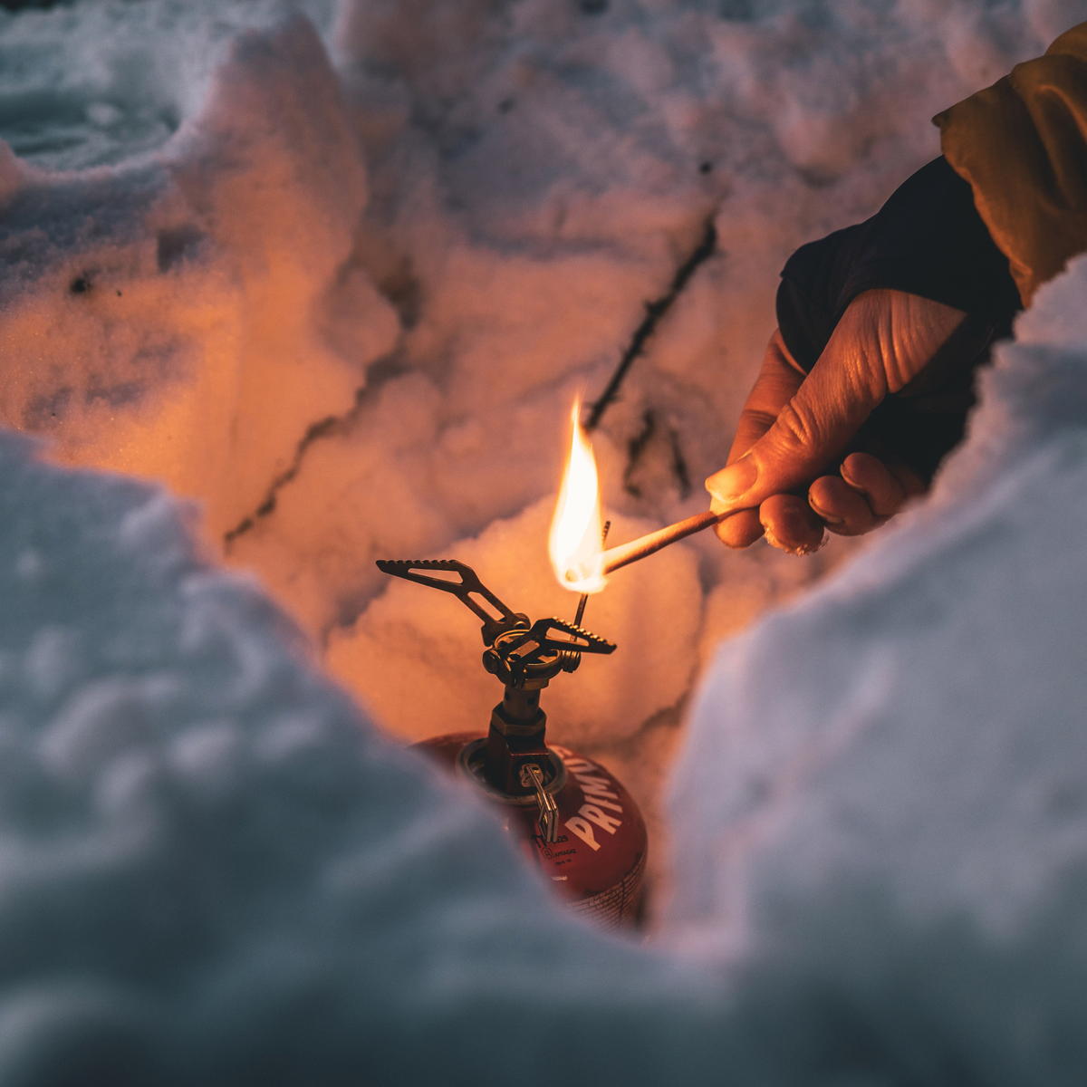 A close-up outdoor shot of a person hand lighting a small camping stove burner with a Lifesystems Stormproof Matches, set in a snowy, icy environment.