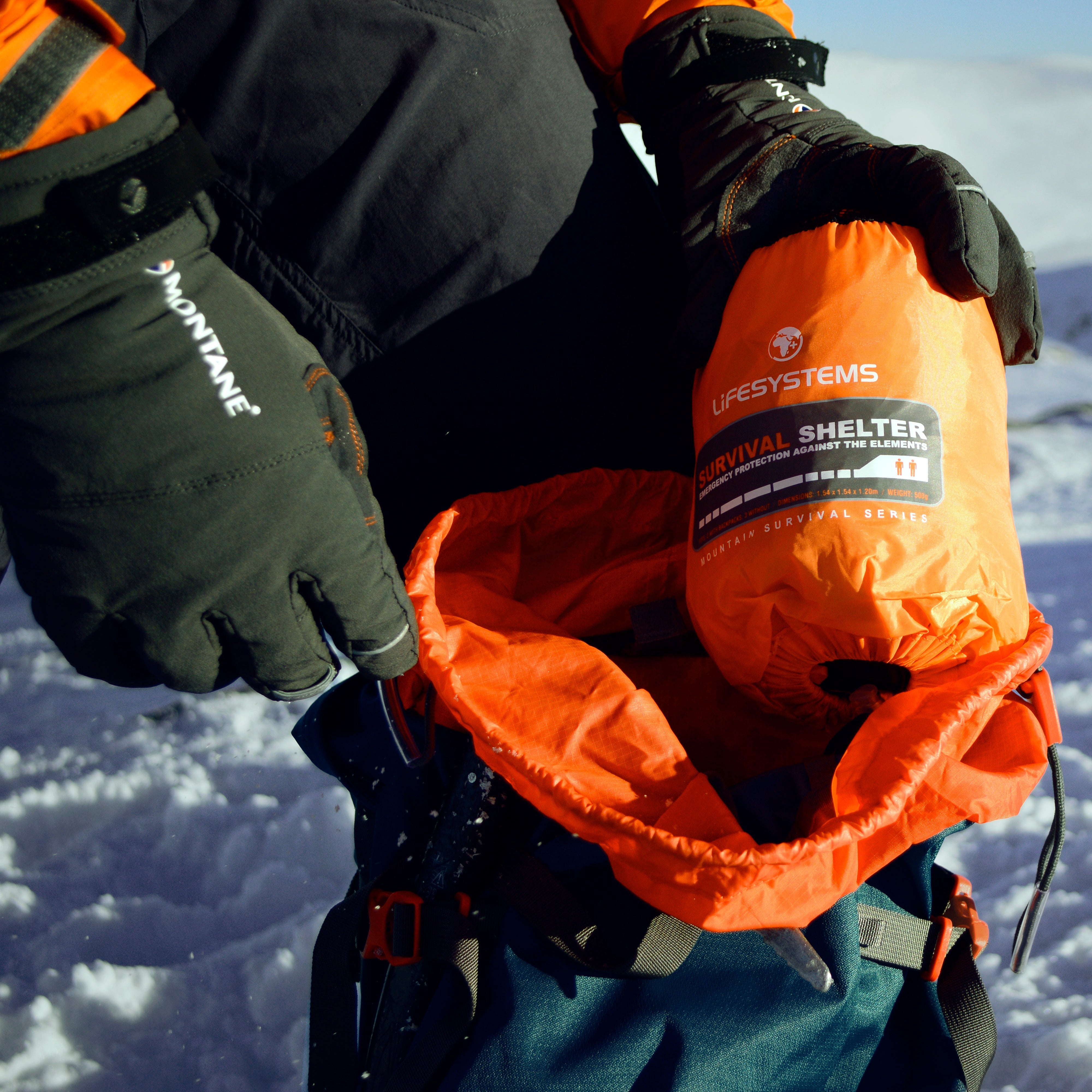 Person wearing gloves taking a Lifesystems Survival Shelter 2 People Orange 42311 out of their backpack with a mountainous snowy landscape in the background