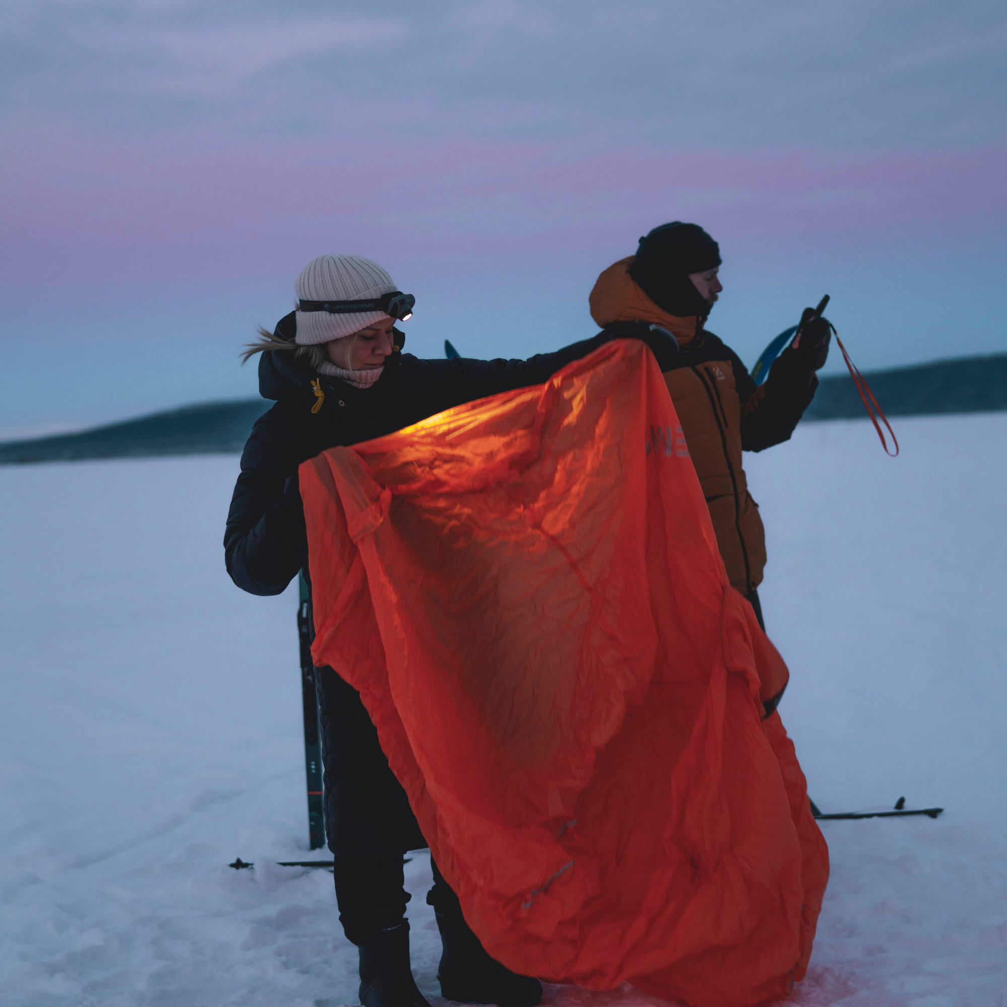 Two people standing on a snowy landscape at dusk, holding up Lifesystems Survival Shelter 4 People Orange 42321. One person wears a white beanie and black jacket, and the other wears an orange puffer jacket.