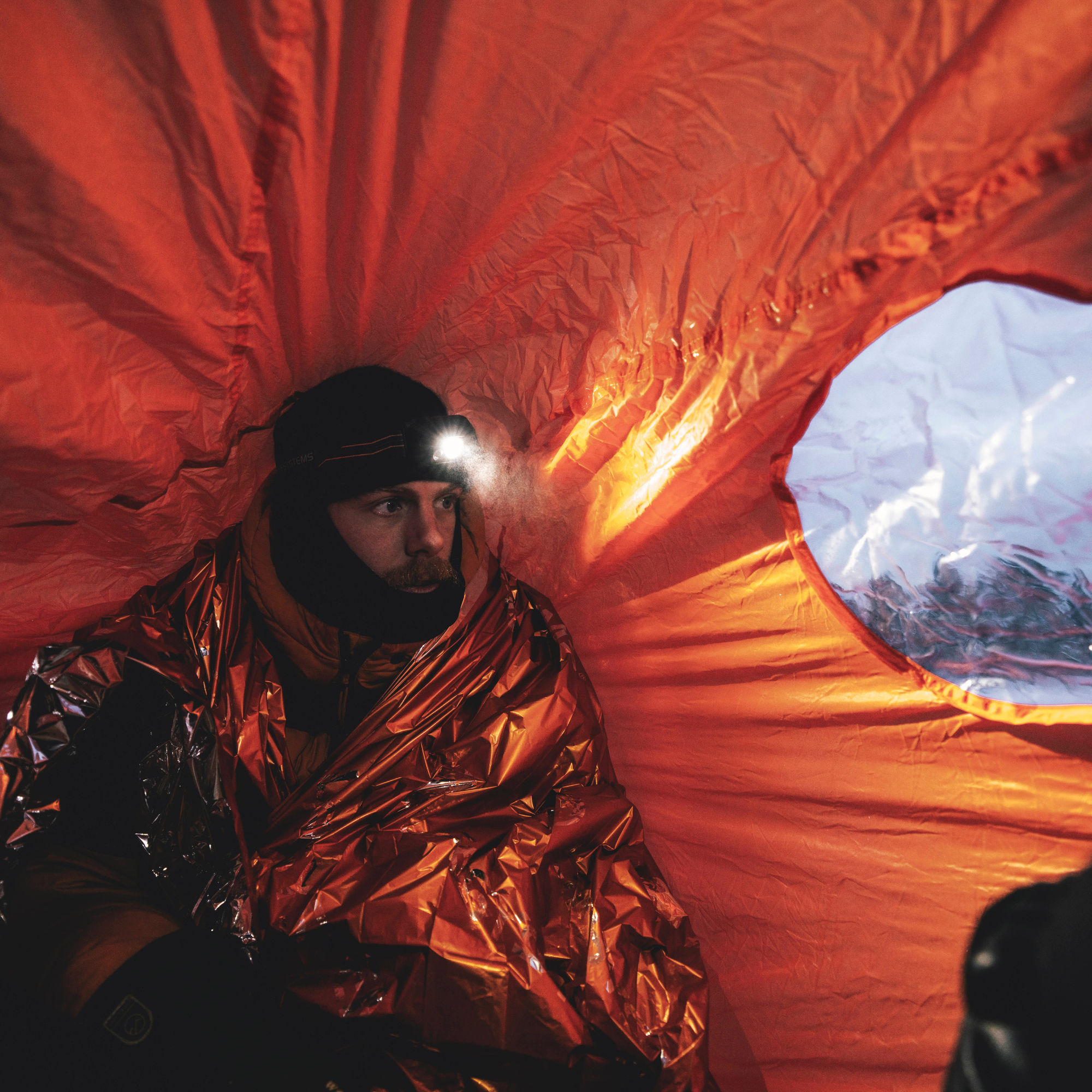 A person inside a Lifesystems Survival Shelter 4 People Orange 42321, wrapped in a reflective emergency blanket, wearing a black beanie with a headlamp on. A circular opening shows a snowy outdoor scene.