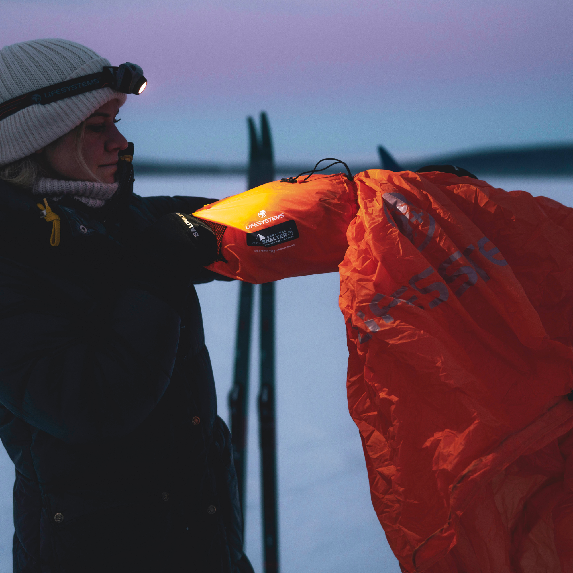 A person outdoors at dusk wearing a white beanie with a headlamp and a dark winter jacket, holding Lifesystems Survival Shelter 4 People Orange 42321. Skis stand in the background in a snowy scene.