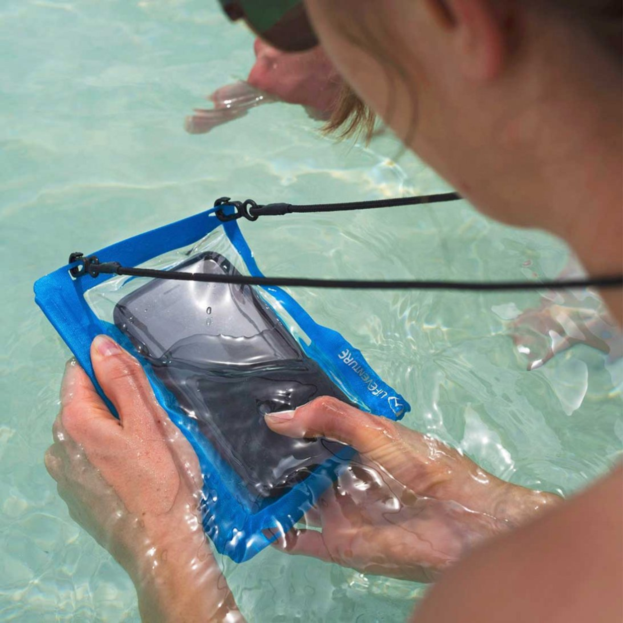 A person in shallow water holds a Lifeventure Hydroseal Waterproof IPX7 Phone Case Blue 59550 with a black phone inside. The pouch has a clear front window and black hanging straps, shown in an outdoor water setting.