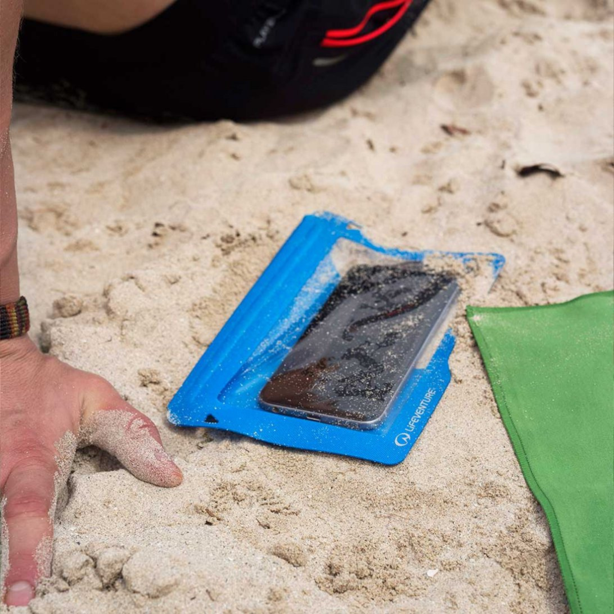 Phone in a Lifeventure Hydroseal Waterproof IPX7 Phone Case Blue 59550 on sand with a hand reaching towards it