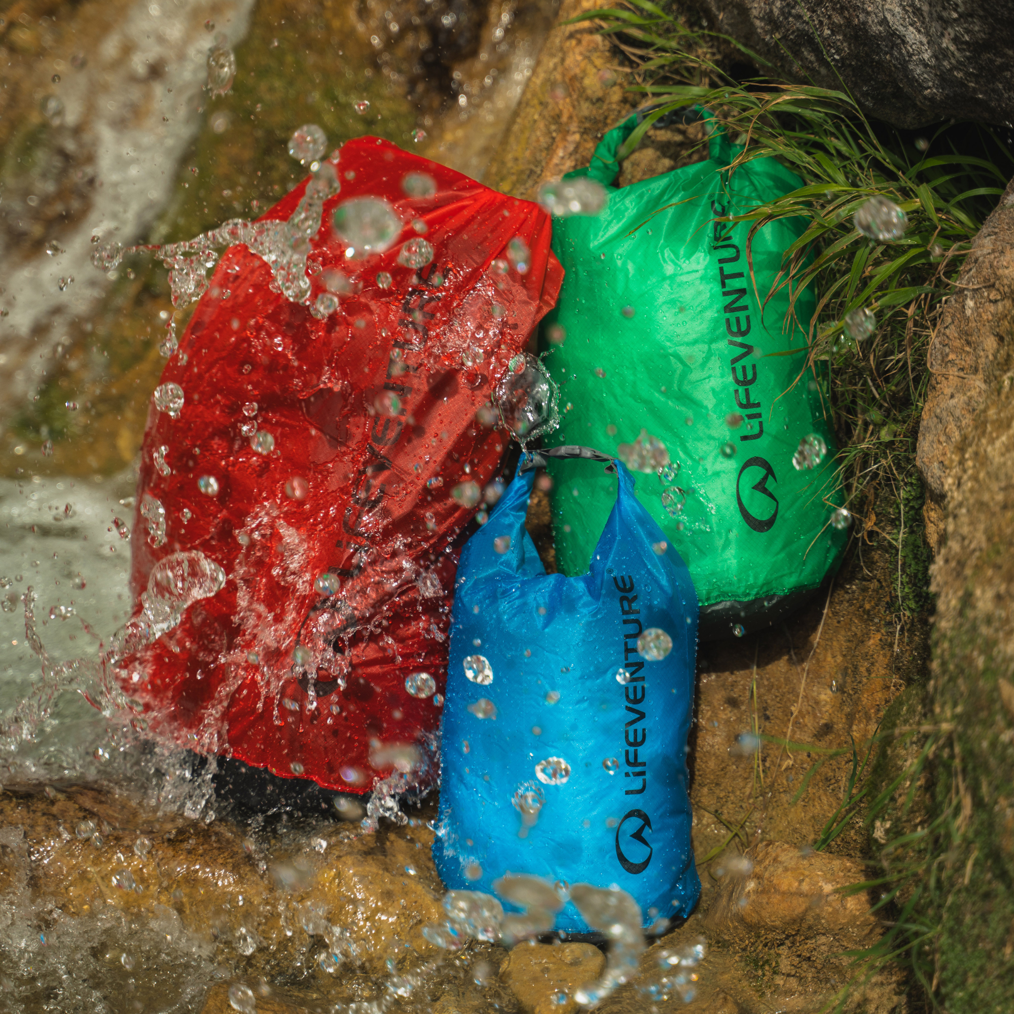 Three waterproof Lifeventure Ultralight Dry Bags in red, green, and blue, shown outdoors in a rocky stream with water splashing around them.