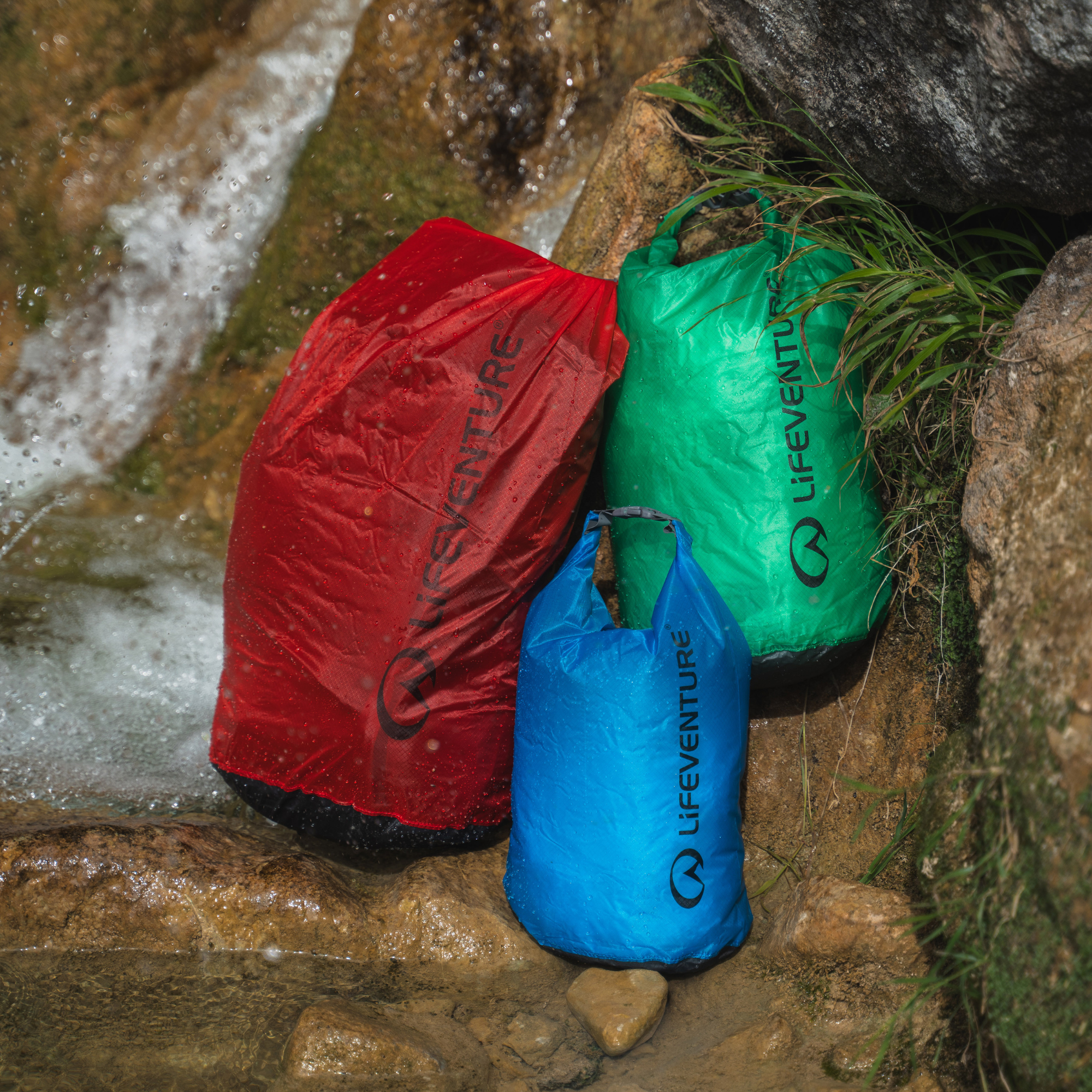 Three waterproof Lifeventure Ultralight Dry Bags in red, green, and blue resting on wet rocks next to a small waterfall.