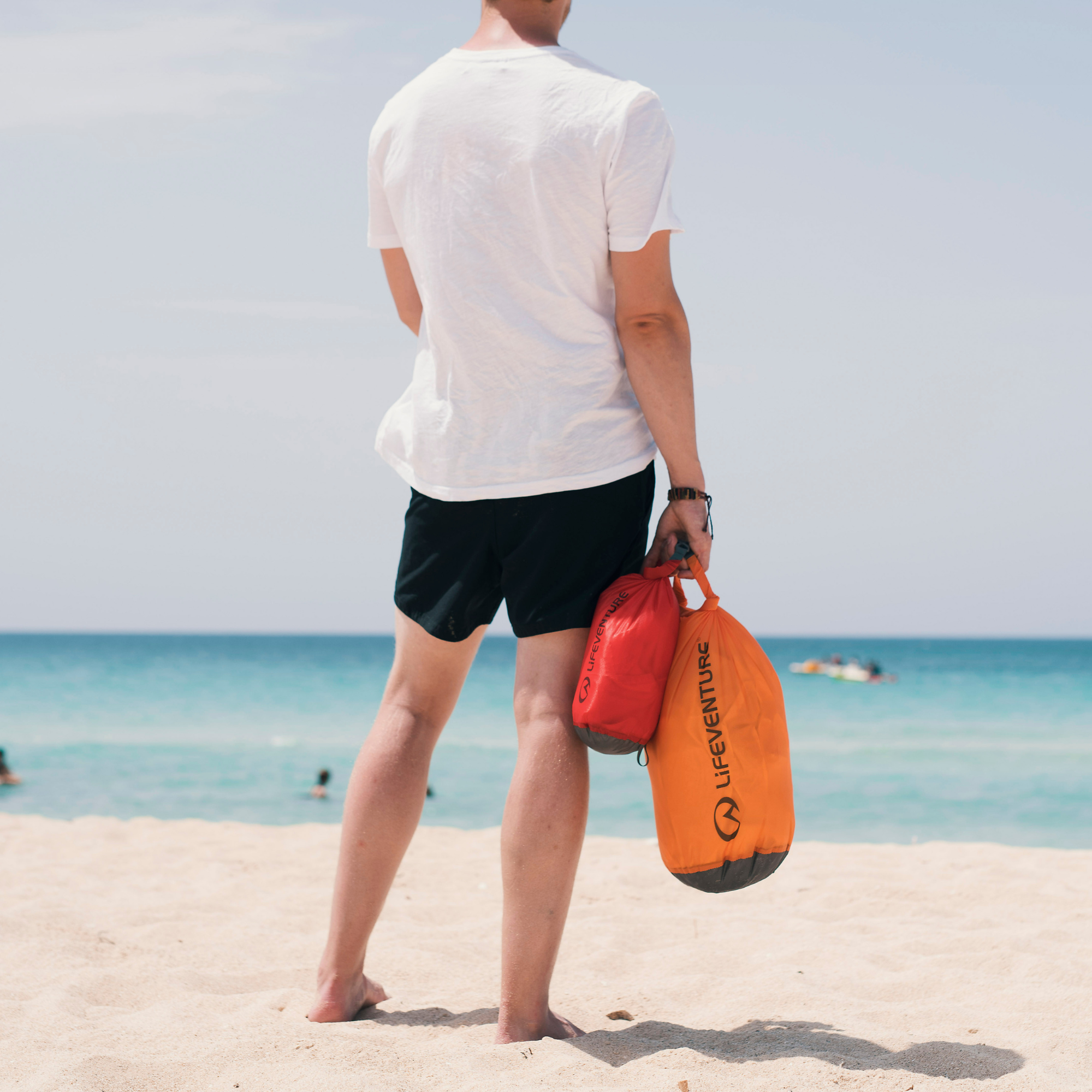 A man standing on a sandy beach facing the ocean, holding two Lifeventure Ultralight Dry Bags, one orange and one red, against a bright beach background.