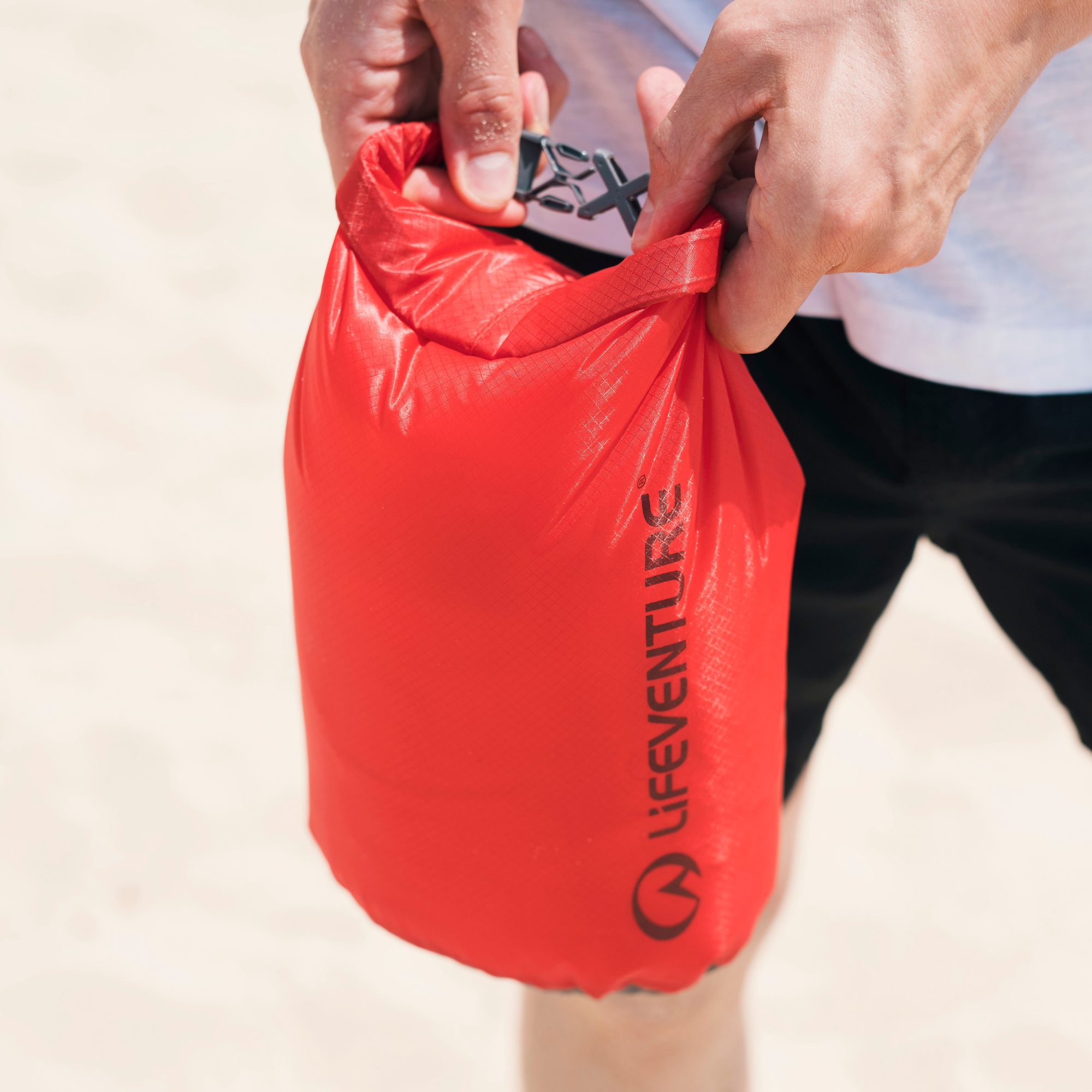 A close-up of a person's hands holding a Lifeventure Ultralight Dry Bag 2L Red 59610 set against a light sandy beach background.