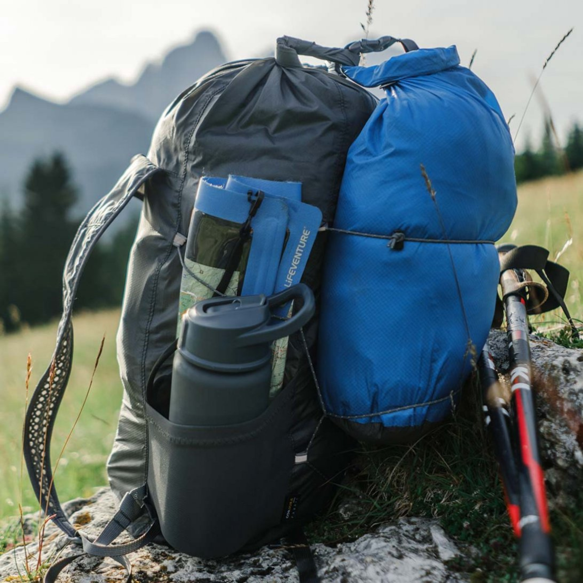 A hiking backpack in dark grey with a Lifeventure Ultralight Dry Bag 5L Blue 59620 attached, set on a rocky grassy hillside with mountains in the background. A trekking pole rests against the rock.