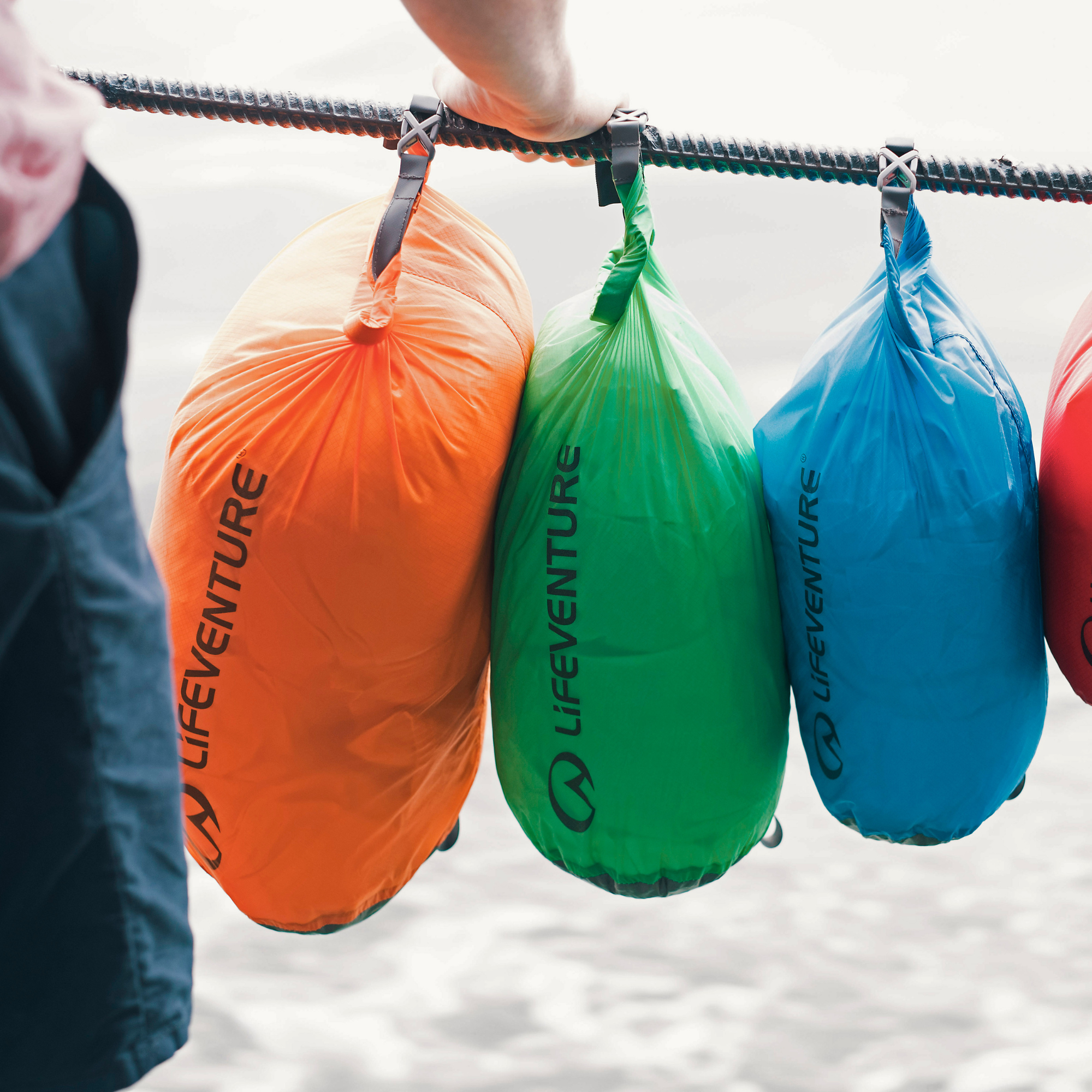 Three Lifeventure ultralight dry bags in orange, green, and blue hanging from a rope outdoors, with a person's hand and leg visible holding the rope.