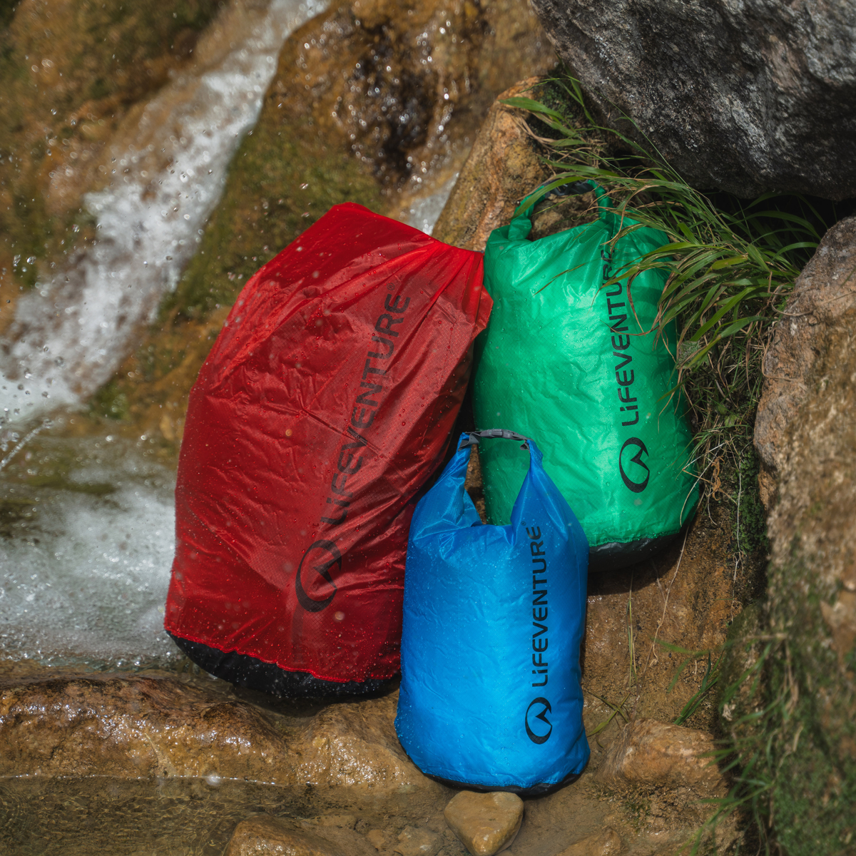 Three Lifeventure Ultralight Dry Bags in red, green, and blue  resting on wet rocks next to a small waterfall.