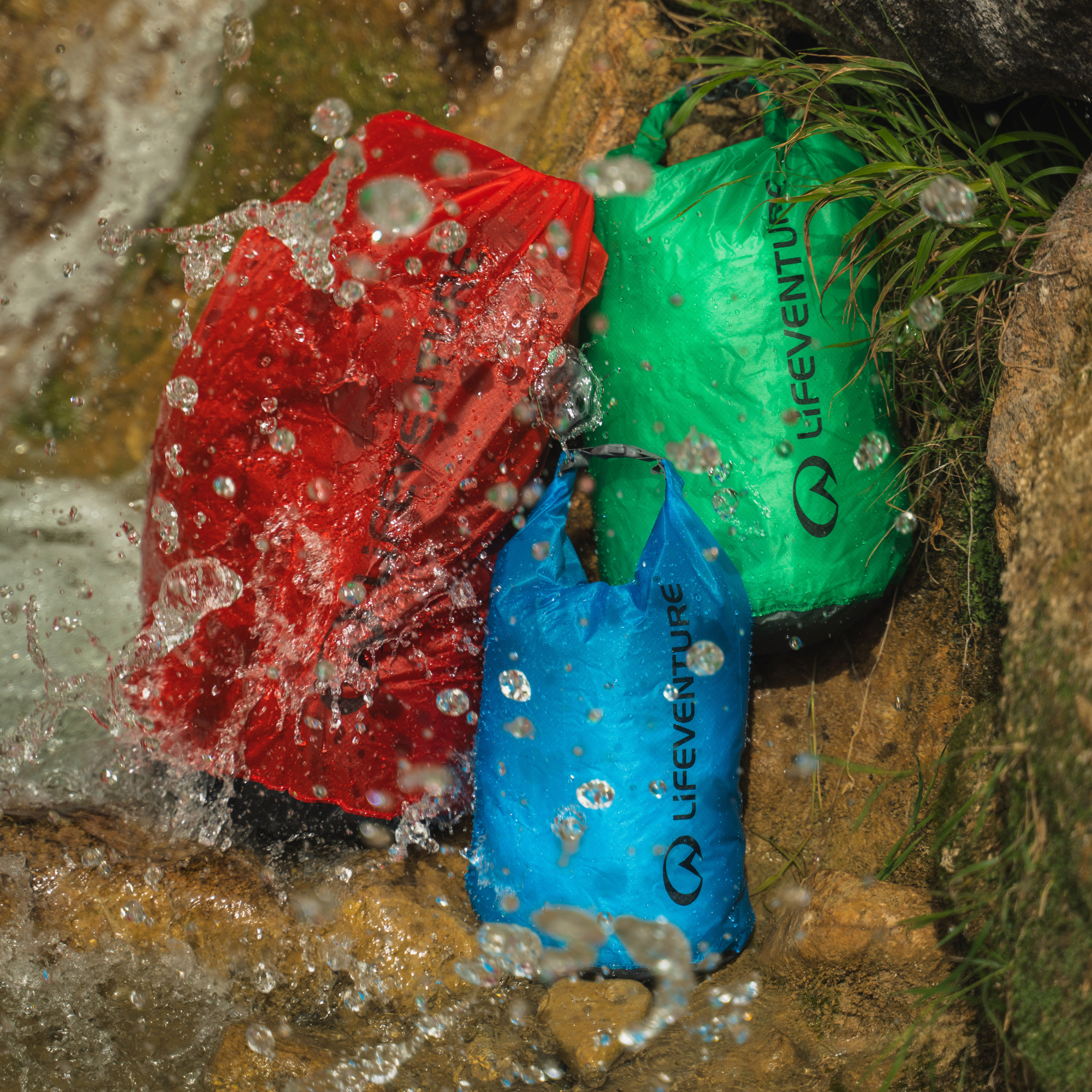 Three Lifeventure Ultralight Dry Bags inred, green, and blue sitting in shallow water with splashing droplets around them.