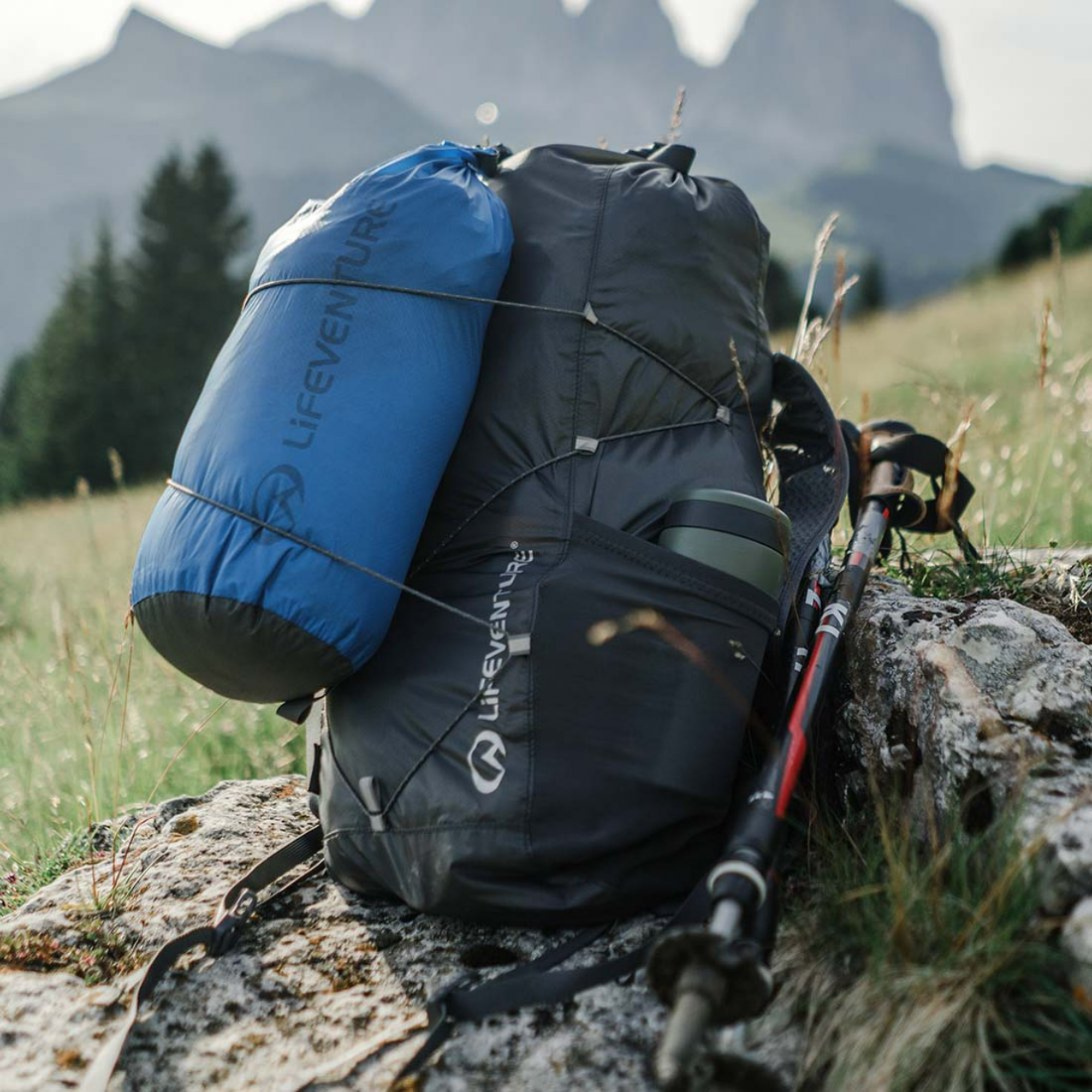 A hiking backpack in dark grey with a Lifeventure Ultralight Dry Bag 5L Blue 59620 attached, set on a rocky grassy hillside with mountains in the background. A trekking pole rests against the pack.