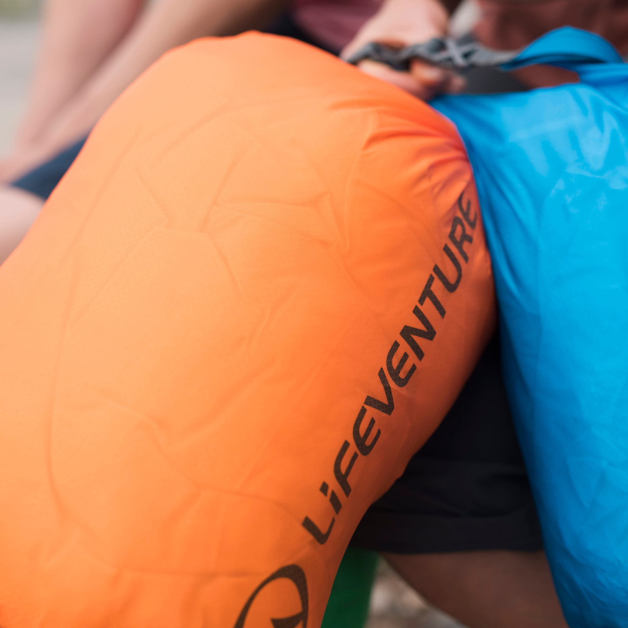Close-up of Lifeventure Ultralight Dry Bag 75L Orange 59680 next to a blue bag, with a person's hands and body partially visible in the background.