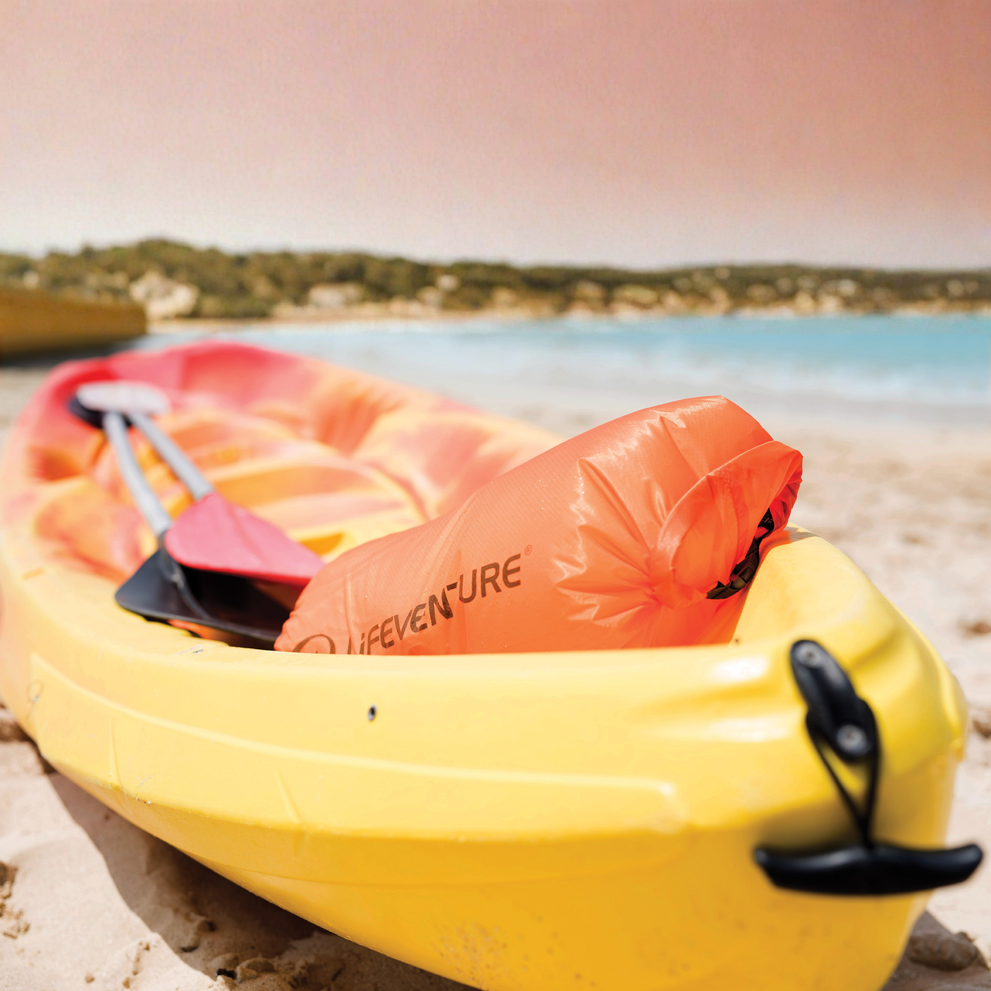 A yellow kayak on a sandy beach with turquoise water in the background. Lifeventure Ultralight Dry Bag 75L Orange 59680 is placed inside the kayak, along with a paddle.