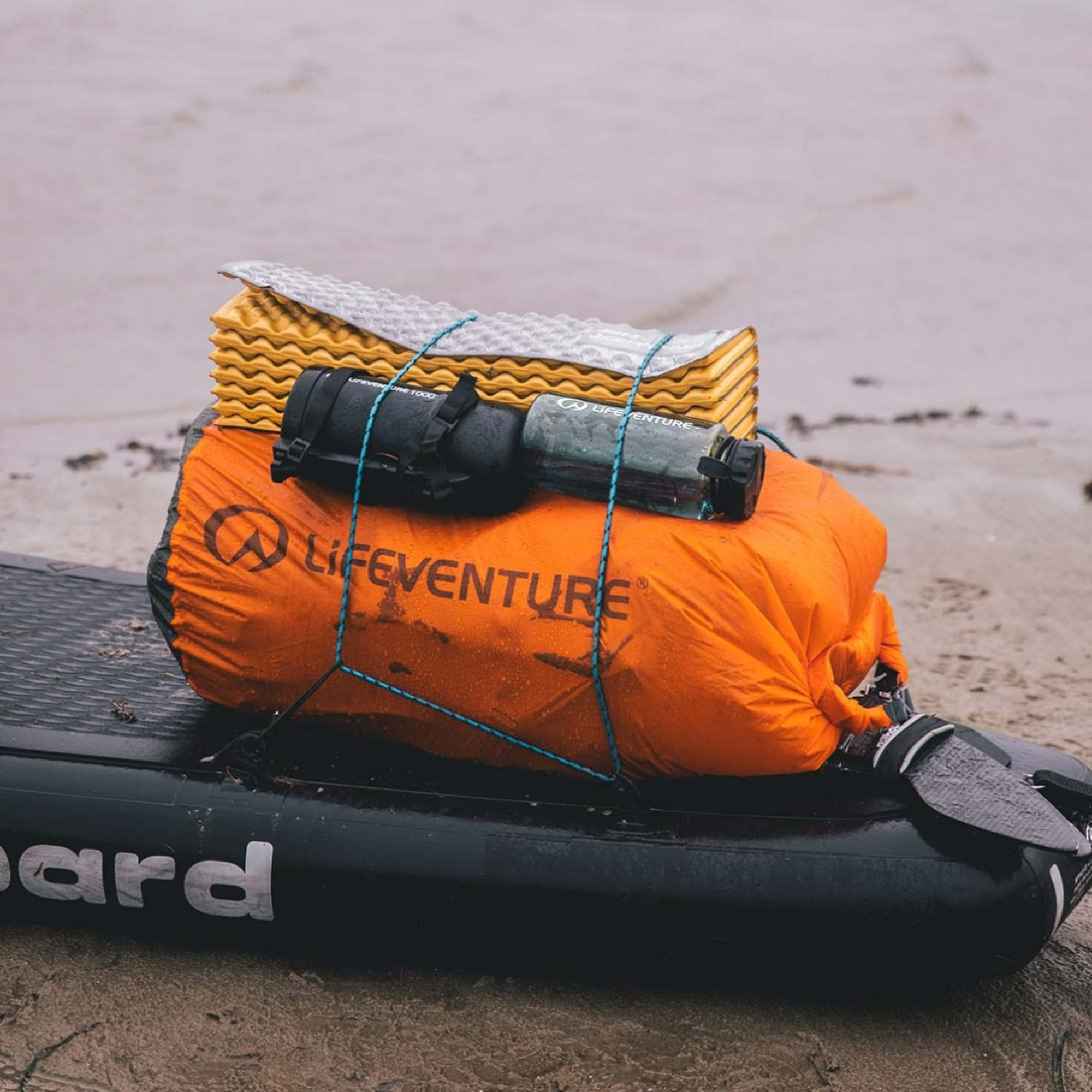 Lifeventure Ultralight Dry Bag 75L Orange 59680 tied to a black stand up paddleboard on a sandy surface