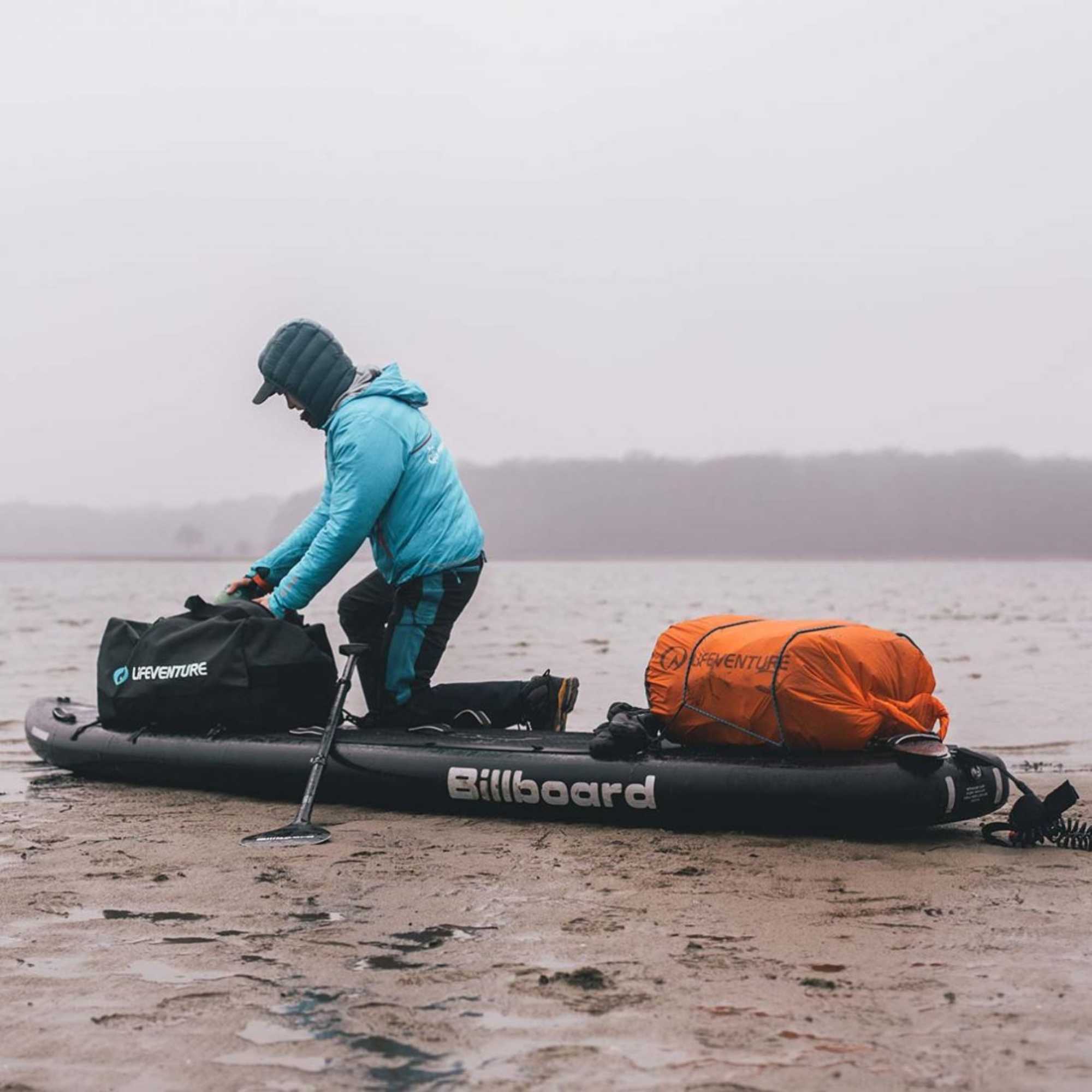 Person preparing a stand up paddle board with equipment on a foggy beach with a Lifeventure Ultralight Dry Bag 75L Orange 59680 strapped to the SUP