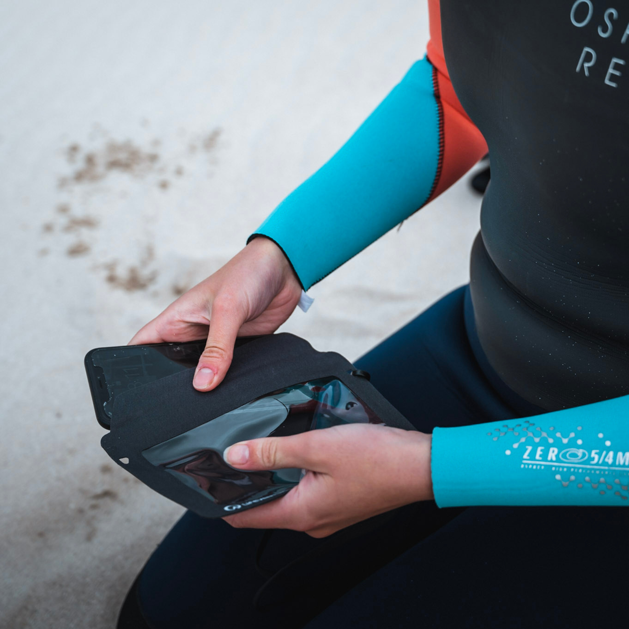 A person wearing a black and turquoise wetsuit placing their phone into an open Lifeventure Waterproof Phone Case Grey 59551 on a sandy beach.
