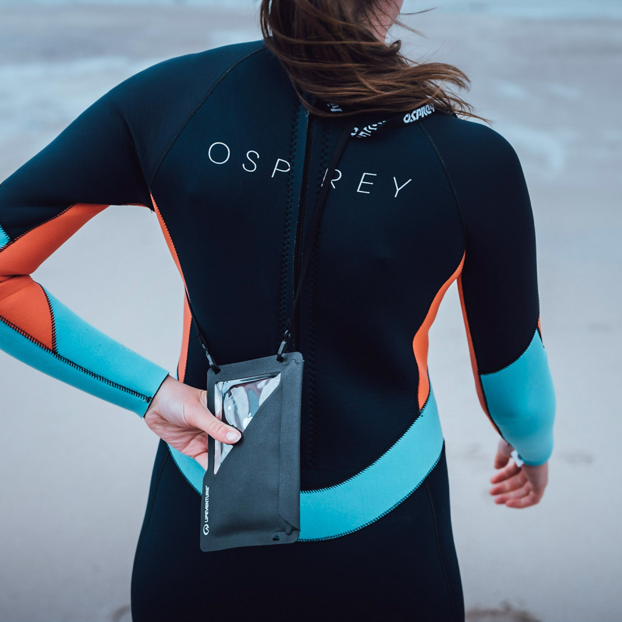 A woman wearing a black, teal, and orange wetsuit seen from behind, holding a Lifeventure Waterproof Phone Case Grey 59551 on a strap around her neck with a soft-focus beach background.