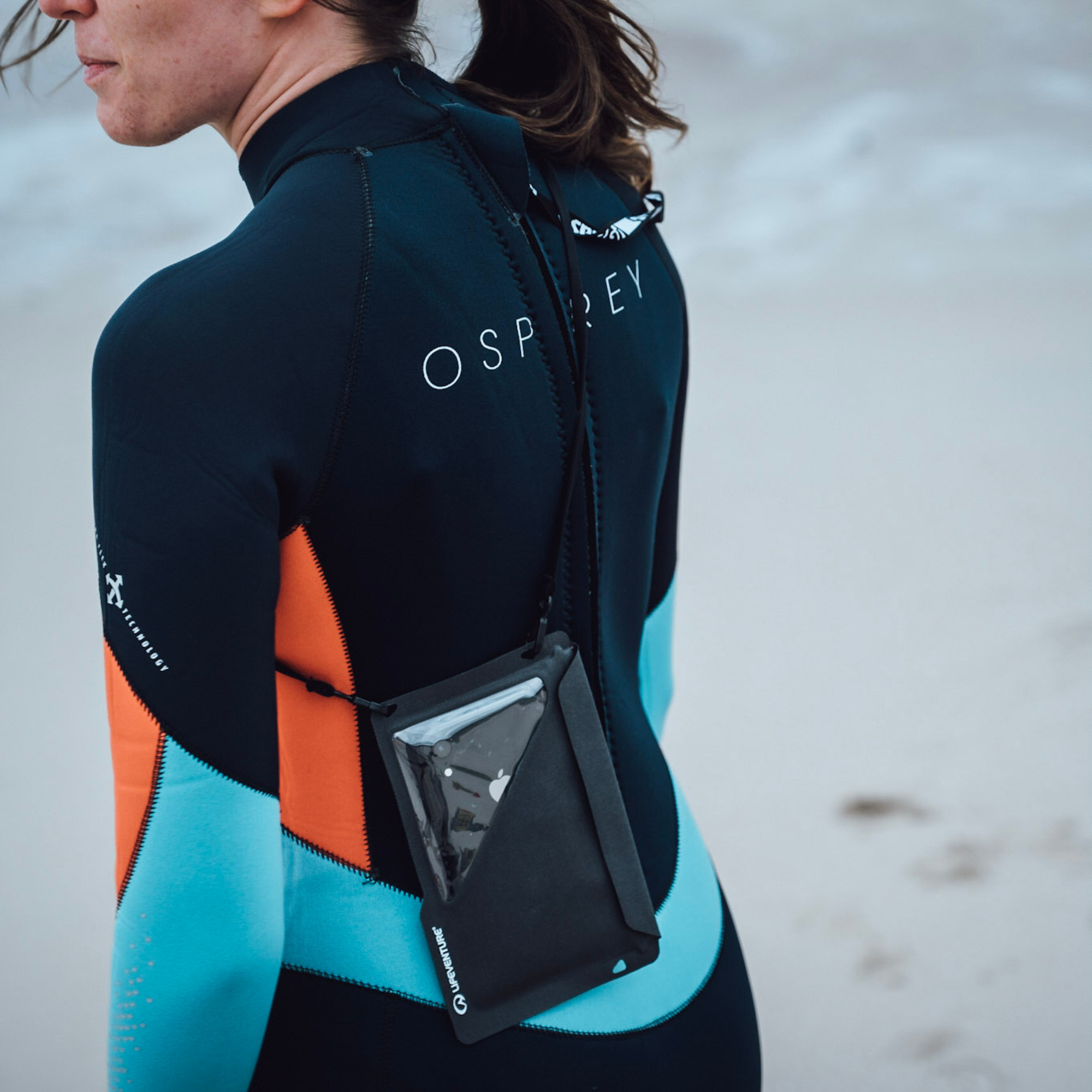 A woman wearing a black, orange, and light blue wetsuit on a beach, carrying a Lifeventure Waterproof Phone Case Grey 59551 across her back.