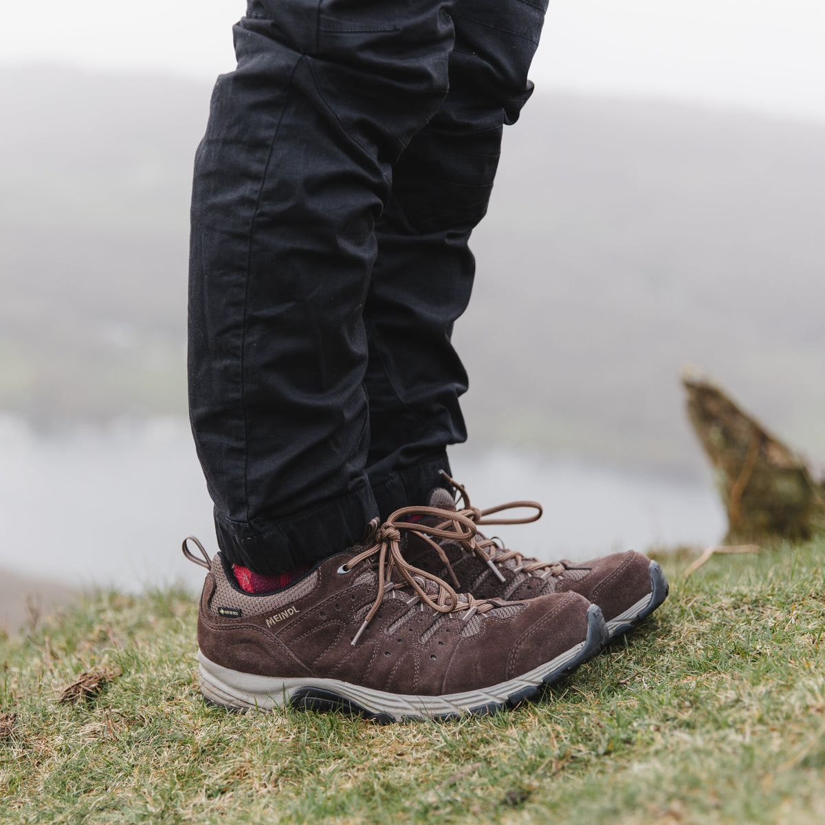 Person wearing Meindl Men's Rapide Gore-Tex Walking Shoes Wide Fit Dark Brown 46 MEINDL5212 and black trousers standing on a grassy area with a blurred natural background