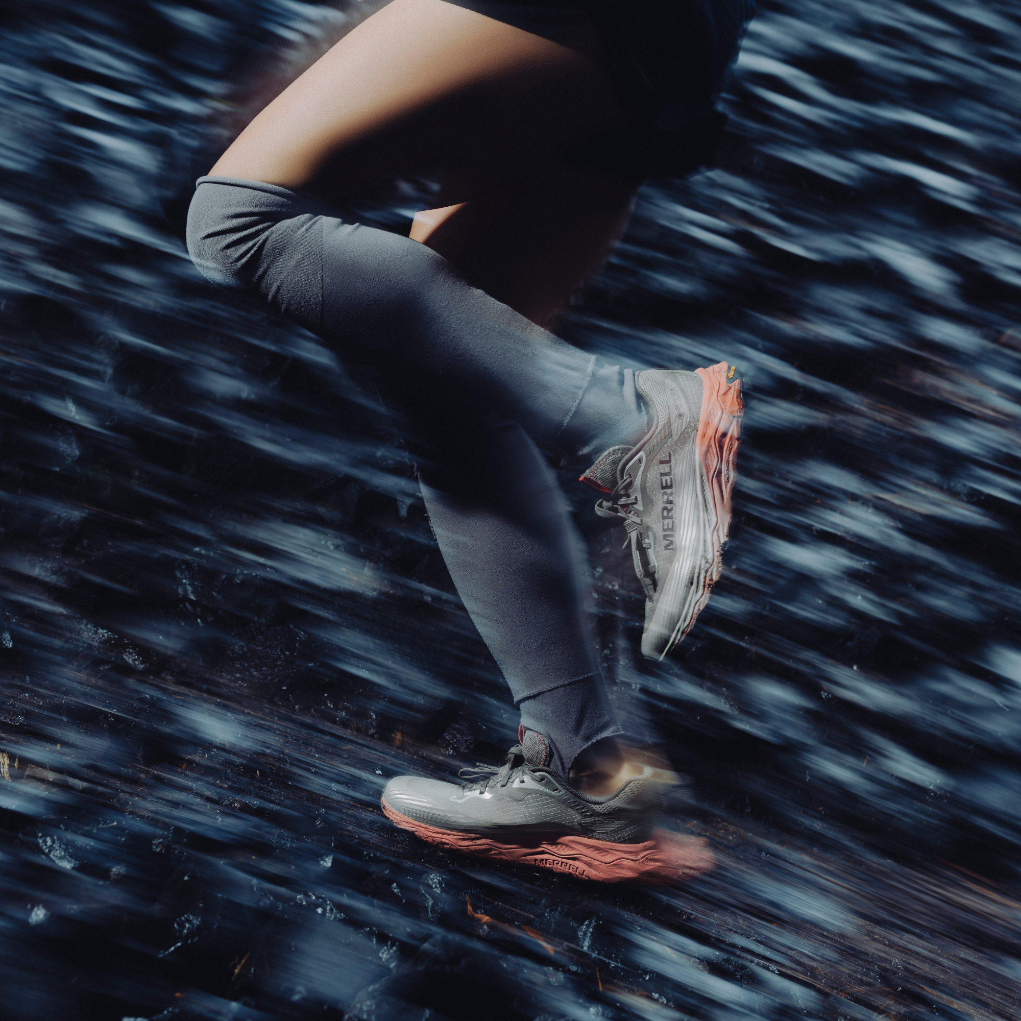 A close-up action shot of a runner's legs in grey compression leggings and Merrell Women's Agility Peak 6 Vegan Gore-Tex Trail Running Shoes Diorite J00005818, captured against a dark, motion-blurred outdoor background.