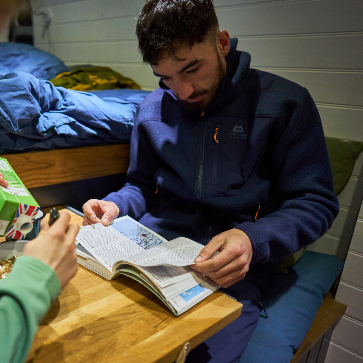 Man wearing Mountain Equipment Men's Highpile Full Zip Fleece Jacket Me-01286 Cosmos ME-009003 reading a trail book sitting in a hostel