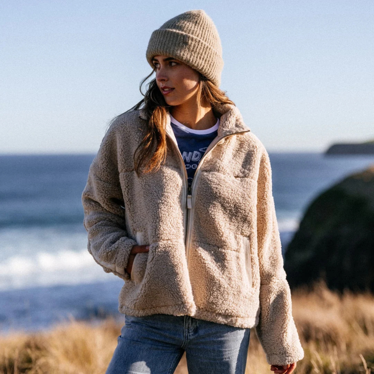 Woman wearing a Rip Curl Women's Cala Nights Sherpa Full Zip Fleece Jacket Natural 0031 02VWJA and beanie standing on a beach.