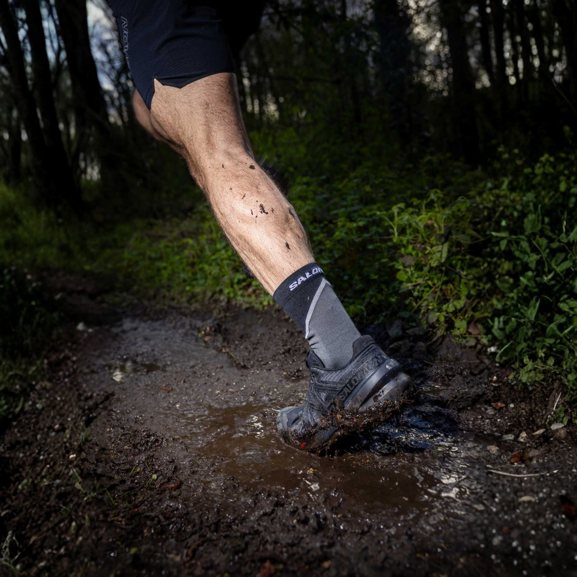 A close-up action shot of a runner's leg kicking up mud on a forest trail, wearing a grey and black Salomon sock and Salomon Men's Speedcross 6 Trail Running Shoes Black Phantom L41737900.