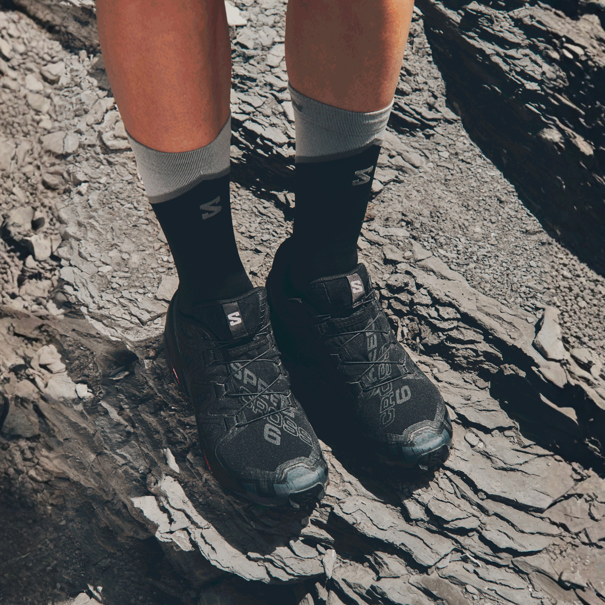A close-up of a person's legs wearing Salomon Men's Speedcross 6 Trail Running Shoes Black Phantom L41737900 and black crew socks while standing on a rocky, outdoor terrain.