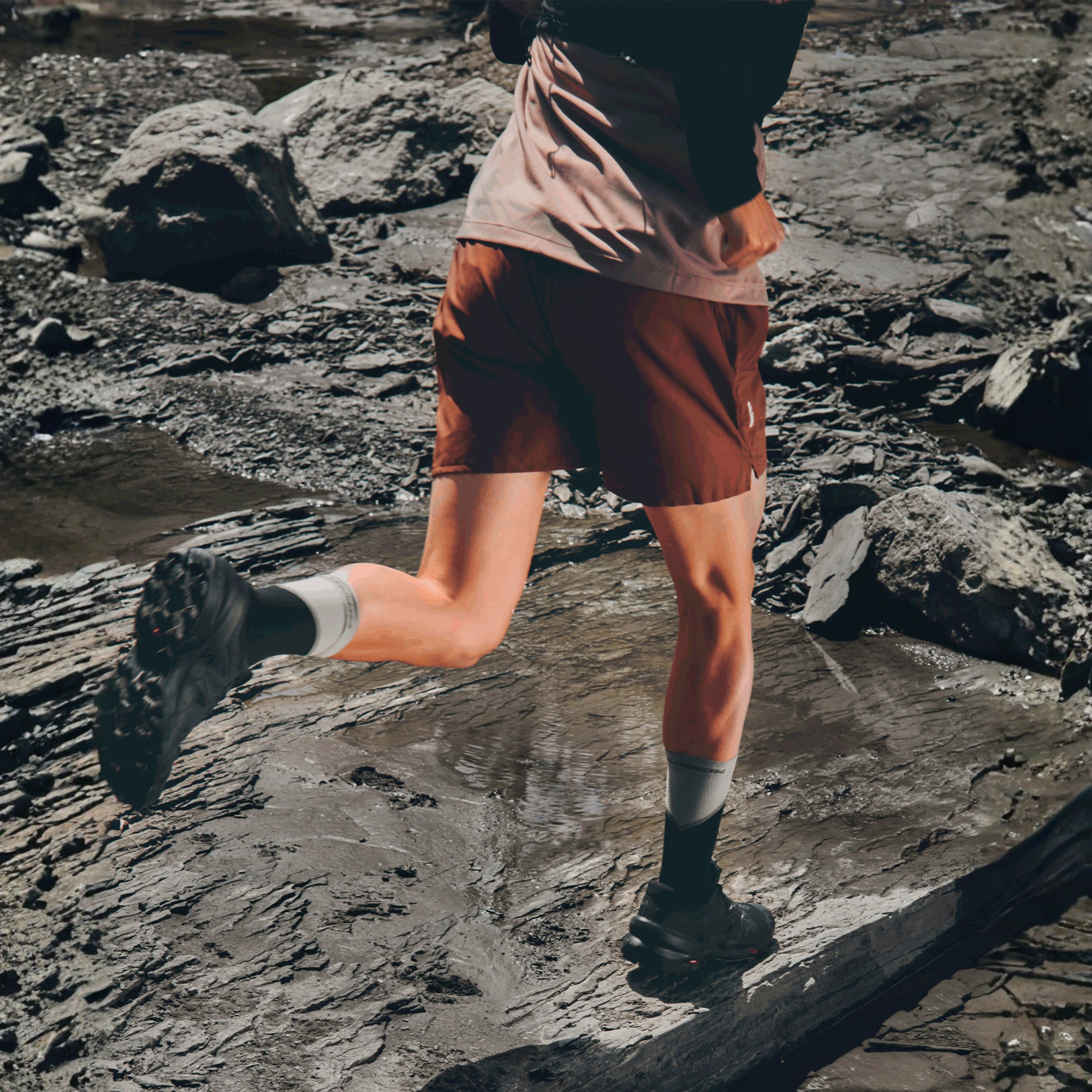 A person running through shallow water on a rocky outdoor trail, shown from the back. The person is wearing Salomon Men's Speedcross 6 Trail Running Shoes Black Phantom L41737900, rust-brown athletic shorts and salmon coloured running top.