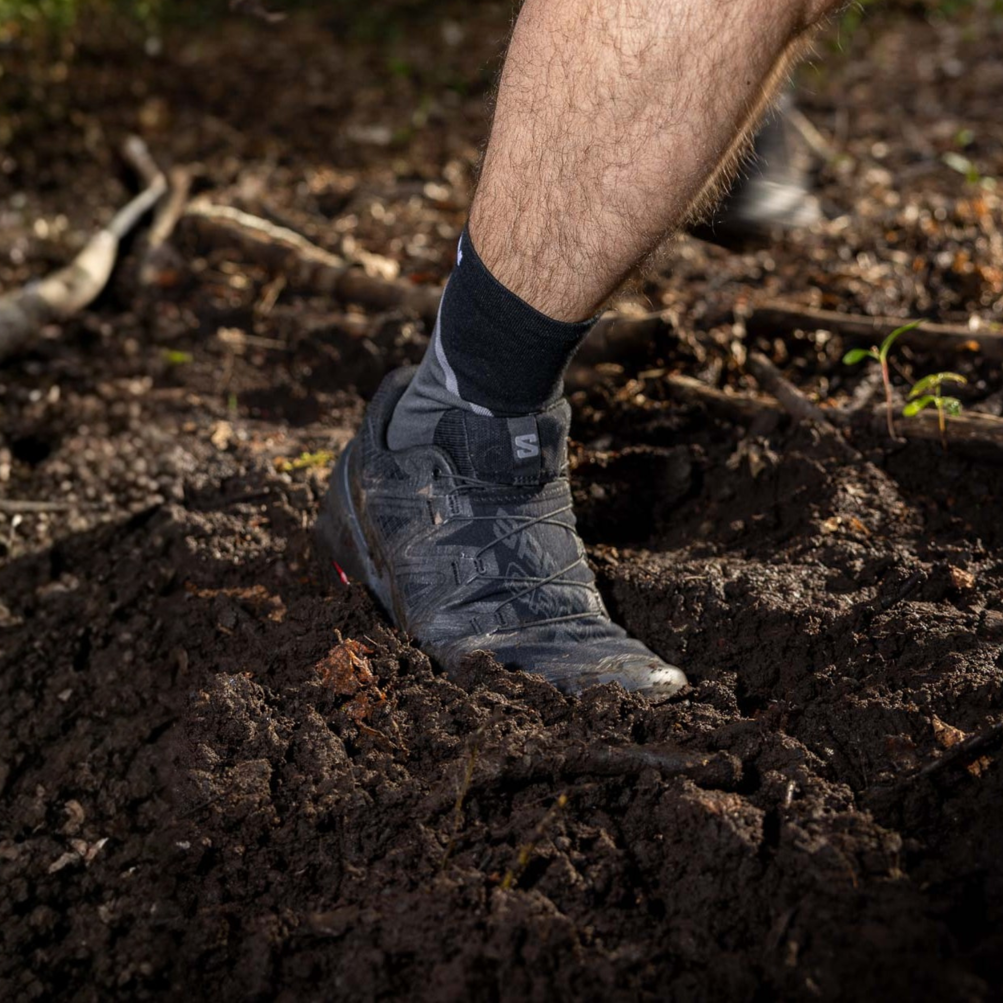 A close-up of a person's lower leg and foot wearing Salomon Men's Speedcross 6 Trail Running Shoes Black Phantom L41737900 and black sock, stuck in deep dark soil outdoors.