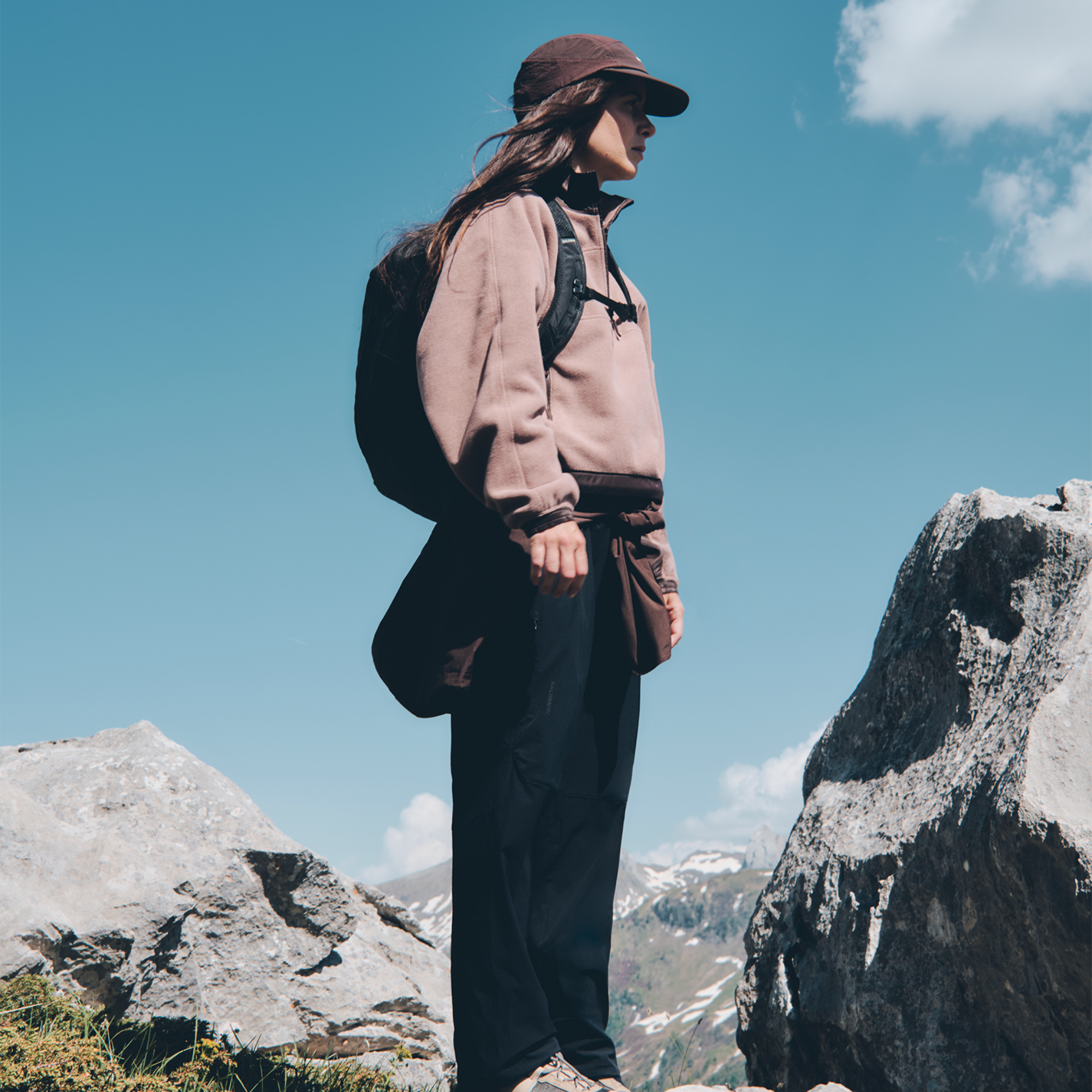 A woman stands outdoors on rocky mountainside under a blue sky, wearing a brown cap, taupe fleece pullover, dark trousers, and a Salomon Trailblazer 20L Daysack Black Alloy LC2182600.