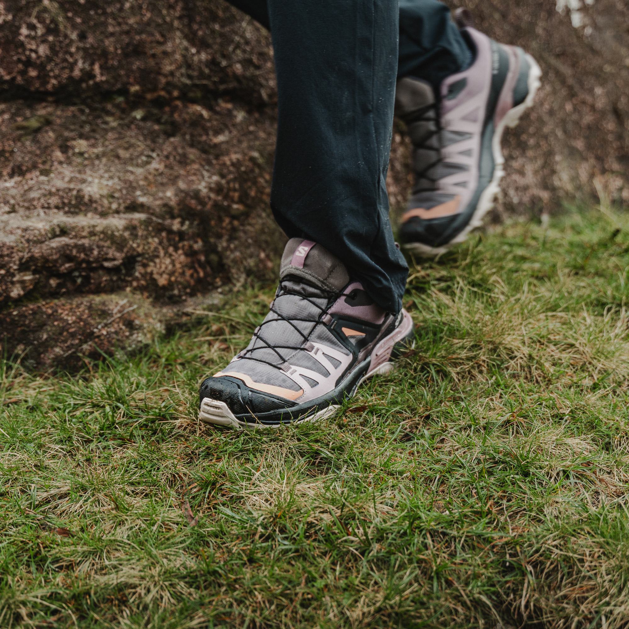 A close-up of a person's legs wearing Salomon Women's X Ultra 360 Gore-Tex Hiking Trail Shoes Plum Kitten Phantom Cork L47449400 and dark trousers, stepping onto green grass next to a large rock.