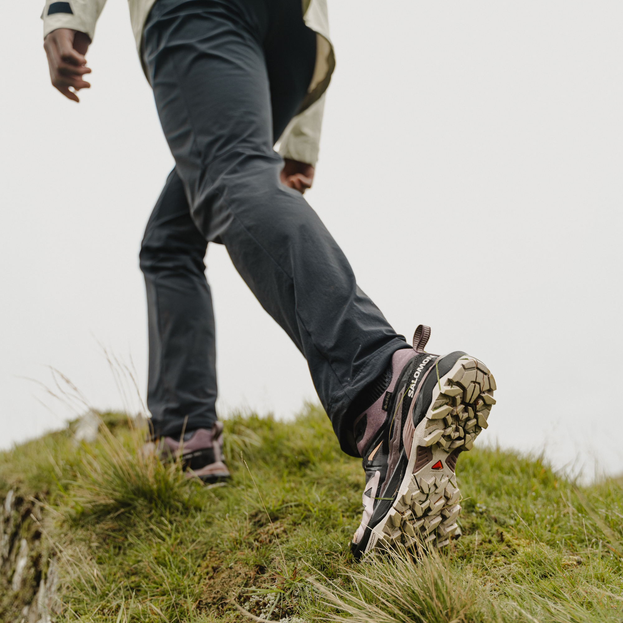 A person hiking up a grassy slope wearing dark grey hiking trousers and Salomon Women's X Ultra 360 Gore-Tex Hiking Trail Shoes Plum Kitten Phantom Cork L47449400.